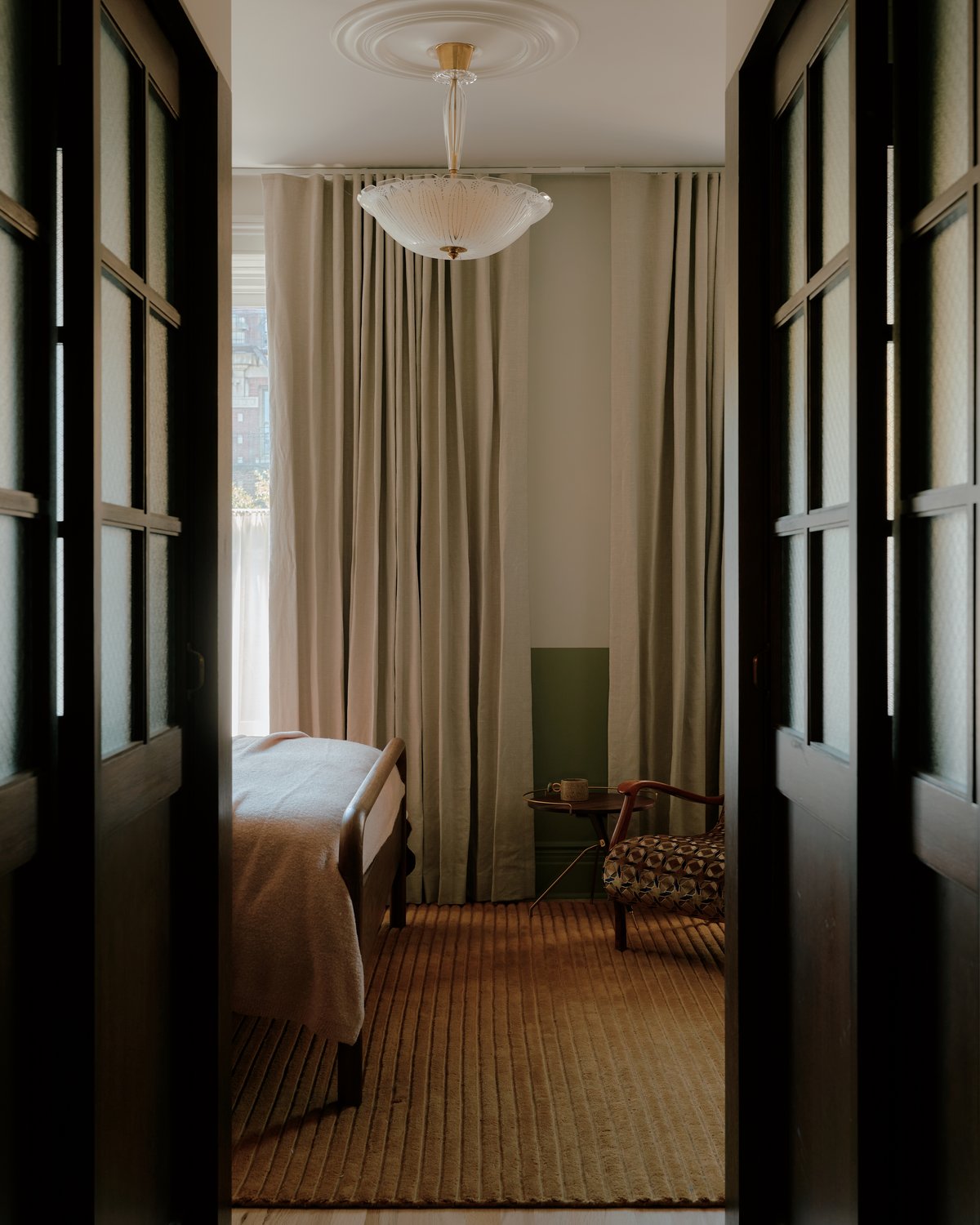 Teppich Park 02 in Camel, in einem Raum mit bodenlangen Vorhängen und einem Bett inszeniert, zeigt einen warmen Kamelton mit subtiler linearer Struktur und niedriger, gleichmäßiger Florhöhe.