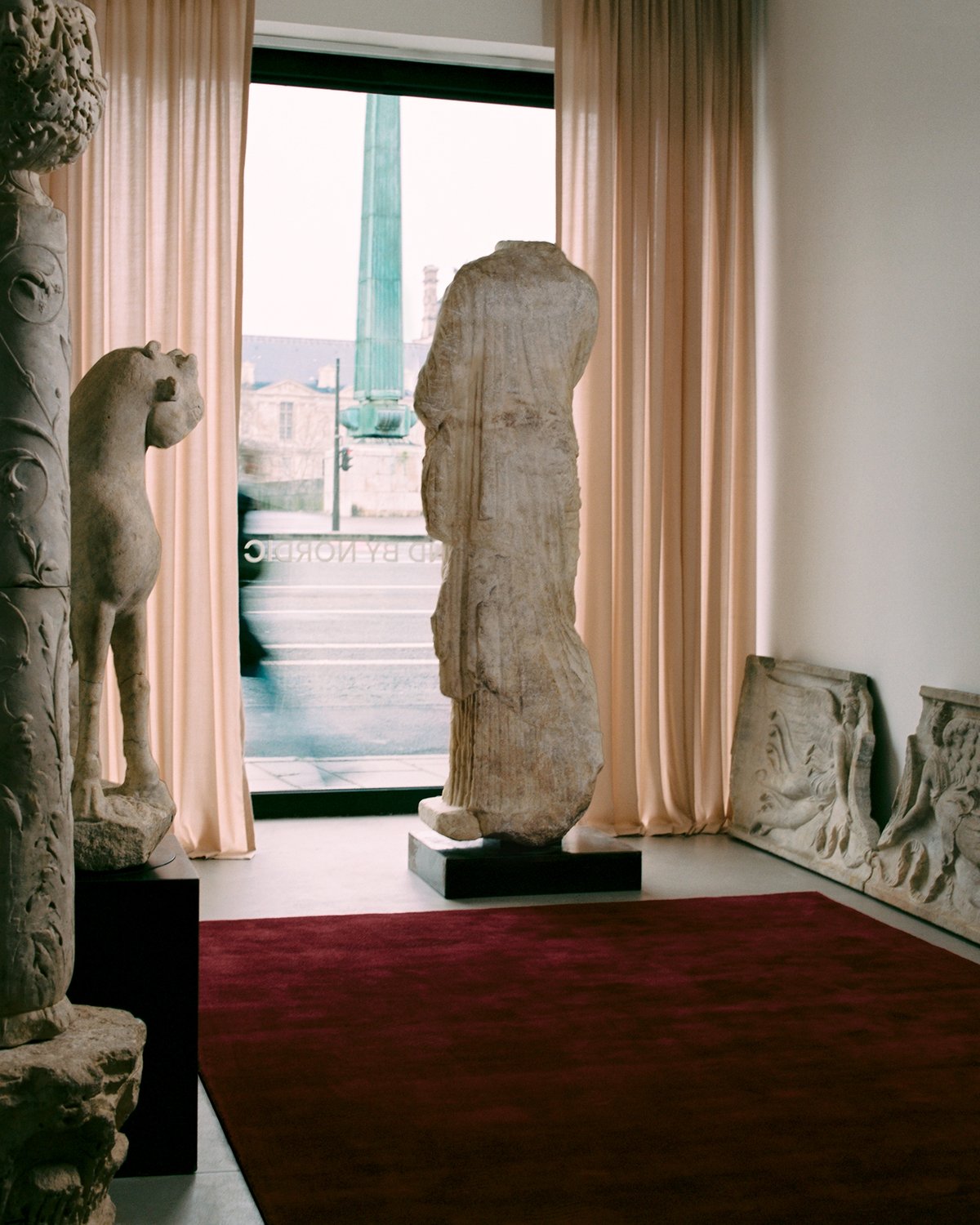 Le tapis Grand en Deep Wine est présenté dans une pièce avec des sculptures, baignée d’une lumière douce filtrée à travers le rideau Grand en Old Rose.