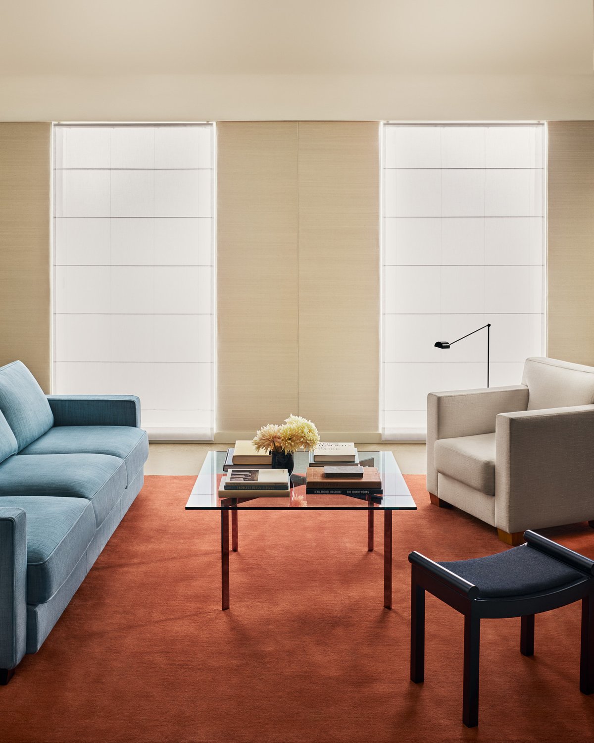 Grand rug in Sienna Red shown in a living room, with a low pile surface and even color, placed beneath a glass coffee table between a blue sofa and a beige armchair.