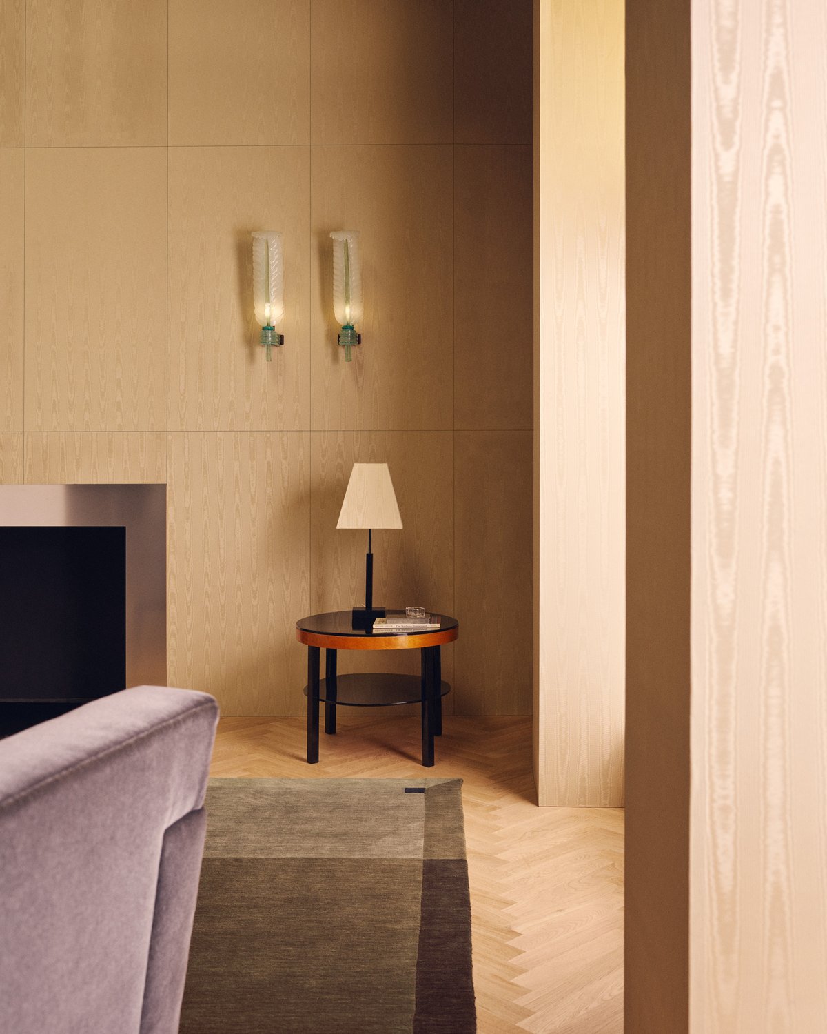 Parchment in Brown, a rug with a tonal border, beside a grey velvet armchair and a round side table with a lamp.