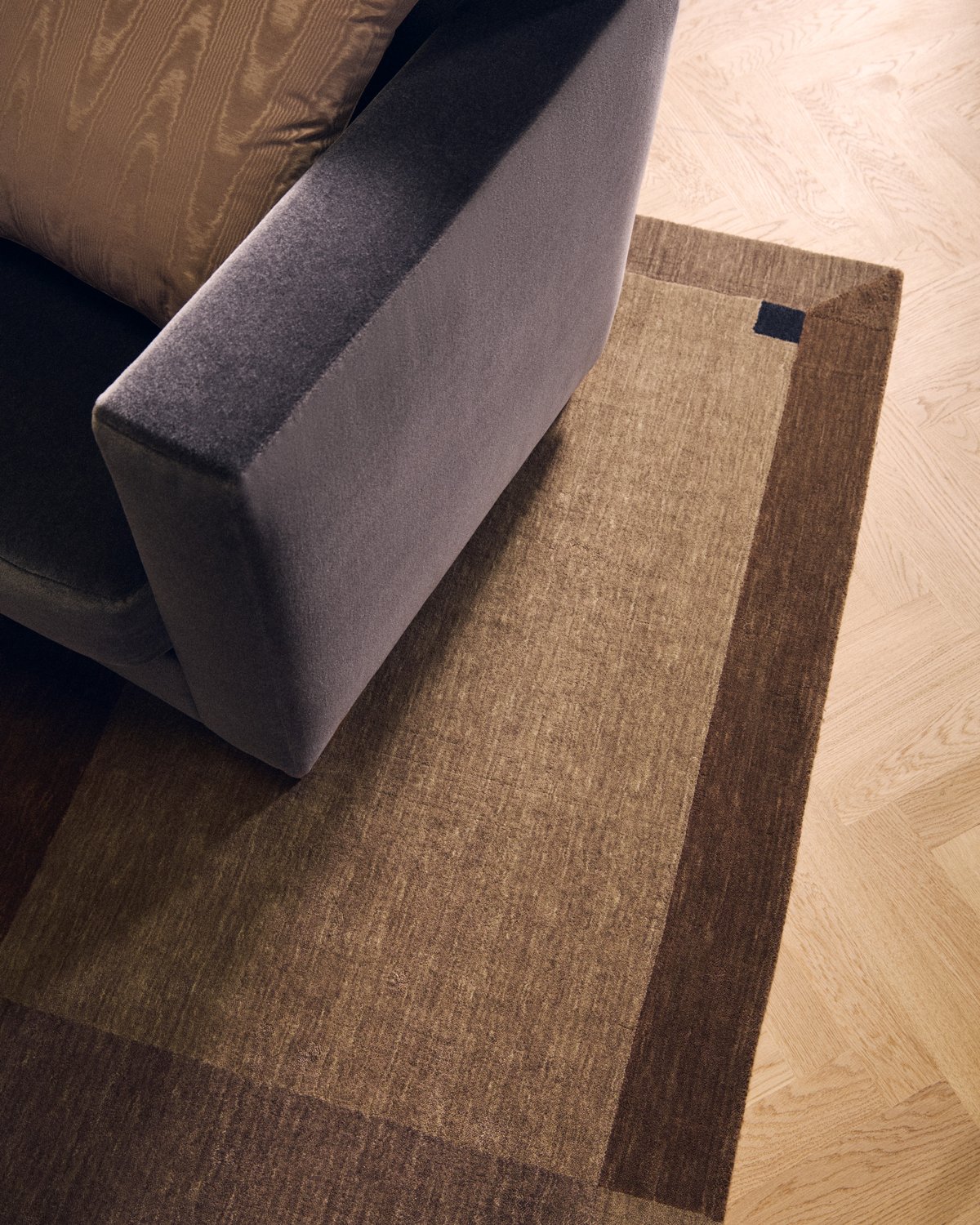 Close-up of Parchment in Brown, a rug with a darker tonal border and a small navy square, beneath a grey velvet armchair.