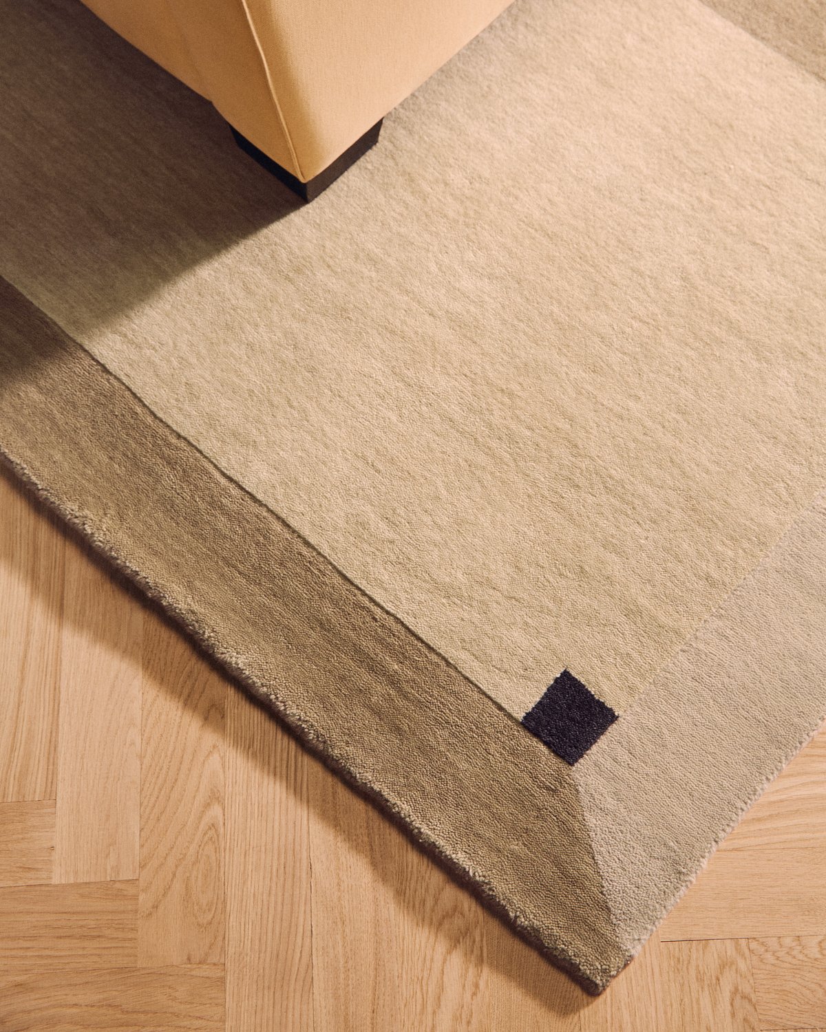 Close-up of Parchment in Sand, a rug with a tonal border and a small dark square at the corner, on a herringbone floor.