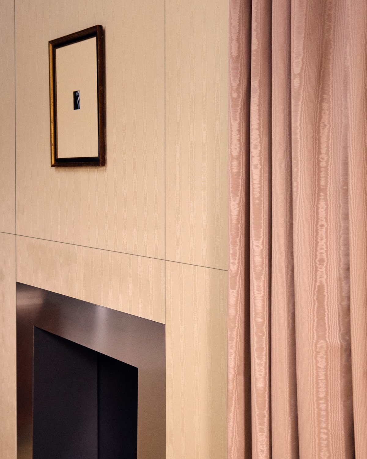 A dusty pink moiré curtain hanging in deep folds beside a moiré-panelled wall and a steel fireplace.