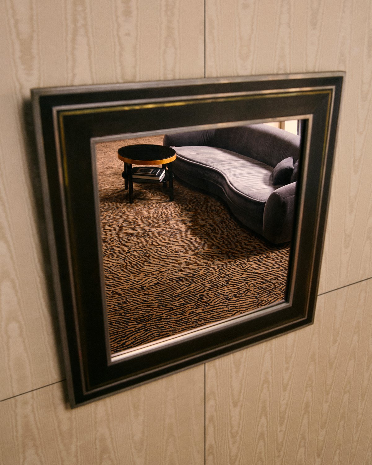 A dark framed mirror on a moiré wall, reflecting a tiger-stripe carpet and a curved gray velvet sofa.