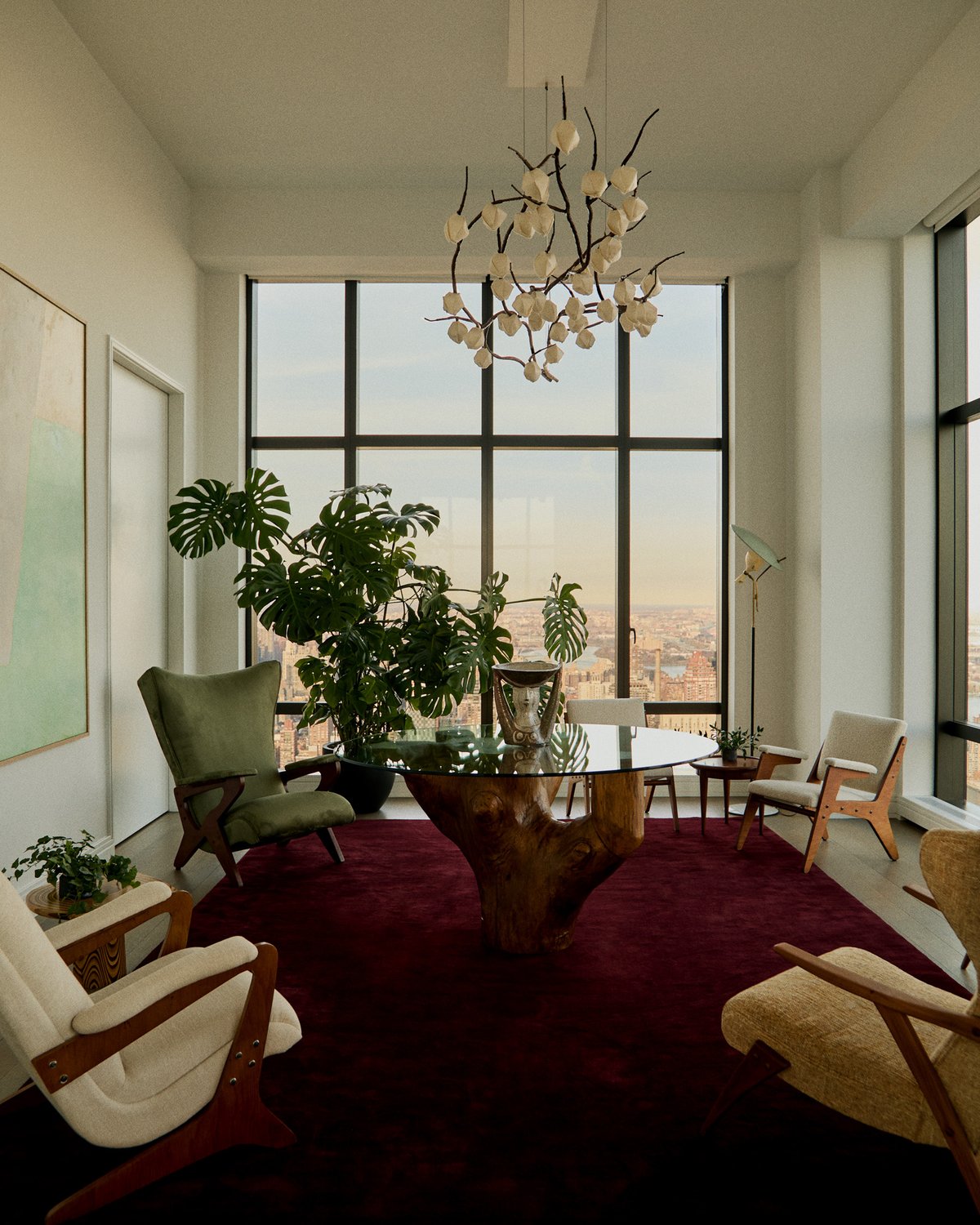 A deep red plush rug beneath a glass table on a tree-trunk base, with armchairs by tall windows over the city.