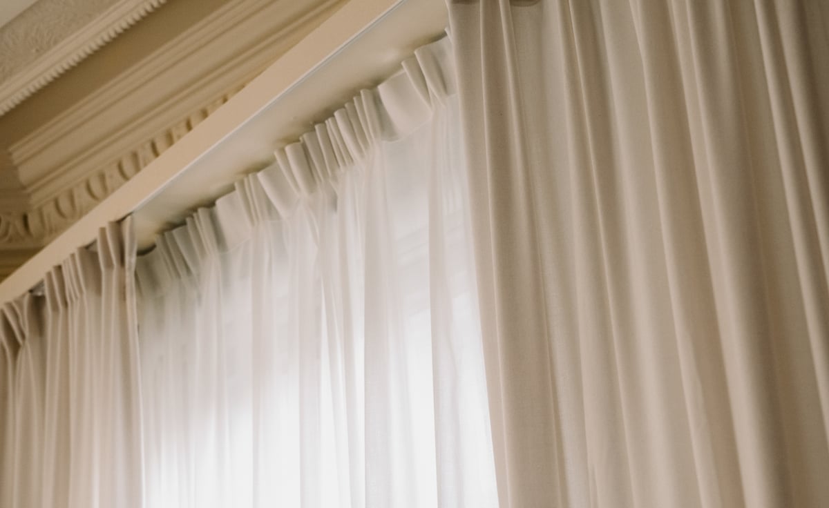 Close-up of The Shade curtain in Warm White layered with The Sheer curtain in Perfect White — a double-layer construction showing a softly textured, opaque fabric paired with a light, translucent sheer, gathered into structured pleats and falling in soft vertical folds.