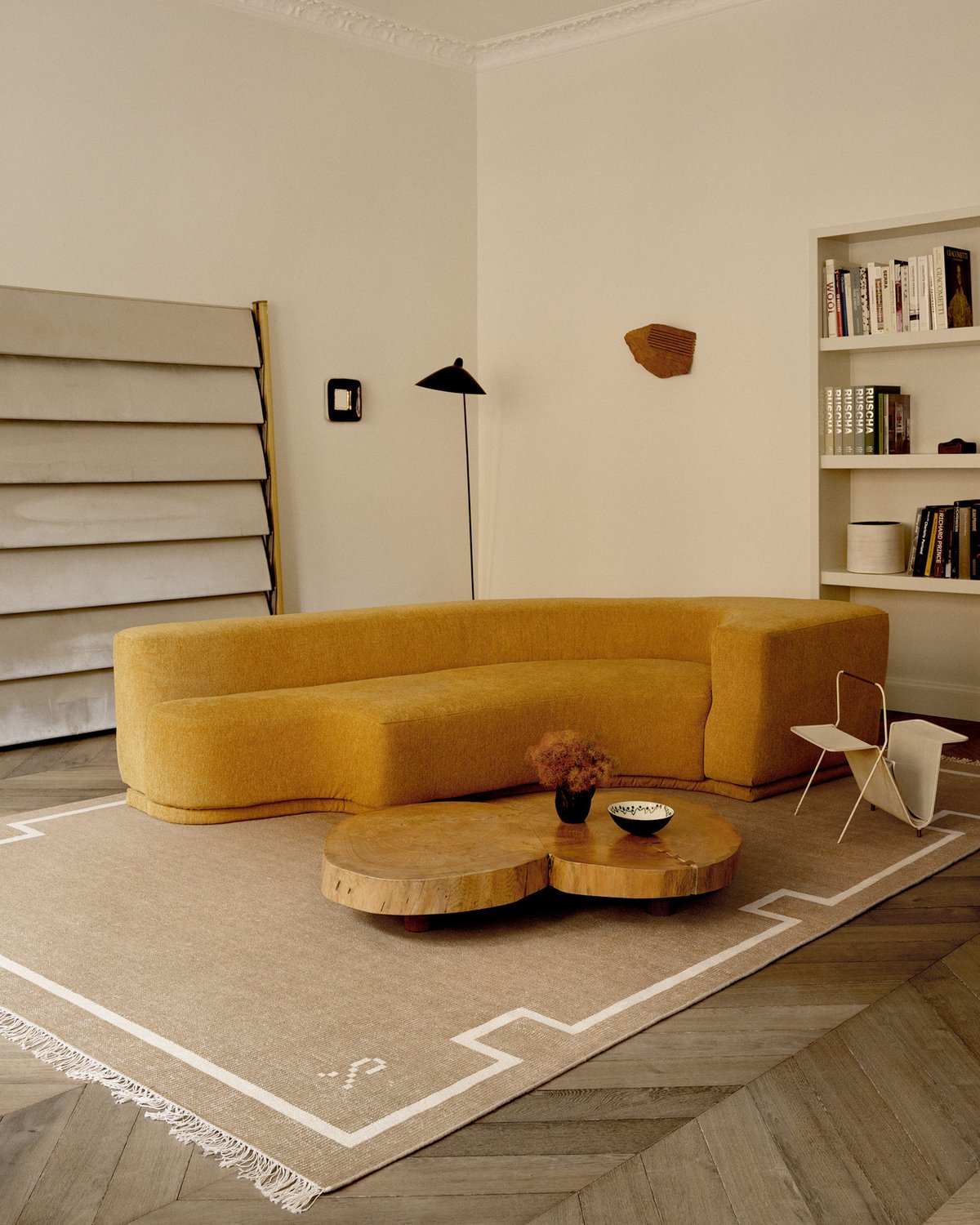 The Elder 05 rug in Cream is shown in a modern living room with a curved mustard sofa, wooden coffee tables, and a geometric floor lamp, creating a warm and minimal atmosphere.