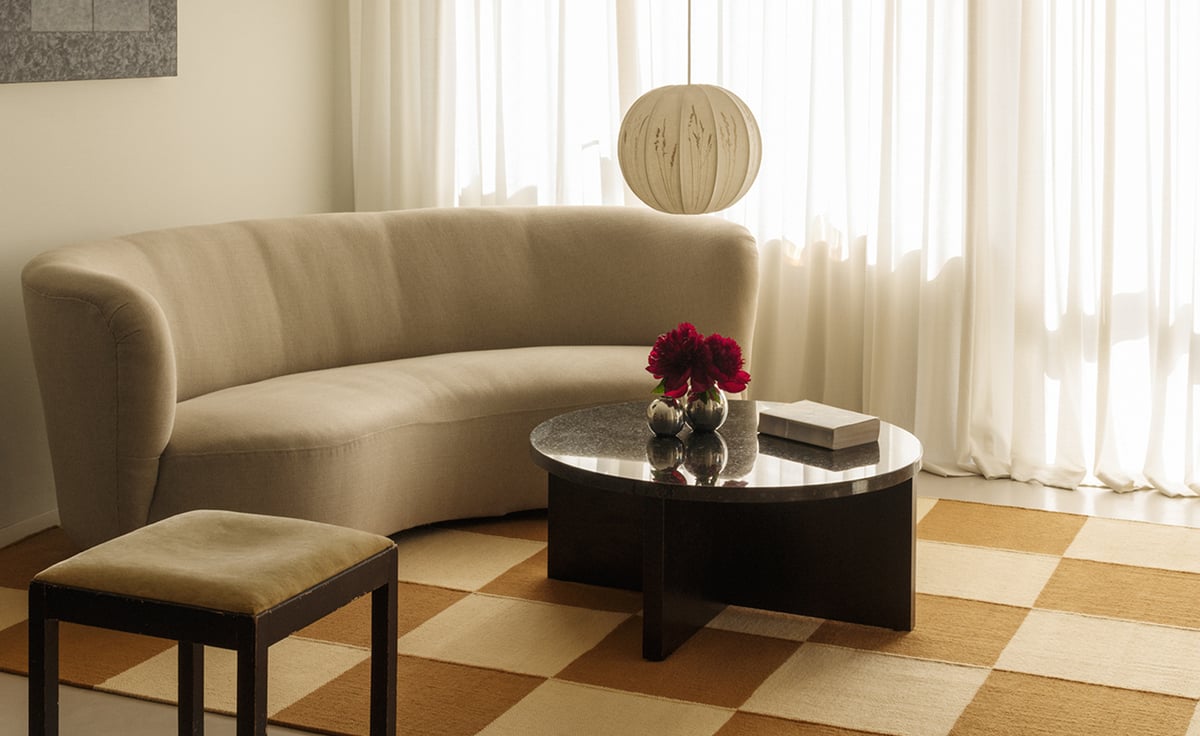 Square – Leo/Yellow sets the tone with its bold checkered design in warm neutrals. A curved cream sofa, sculptural black stone coffee table, and vintage wood-framed stool bring balance and softness to the graphic grid, while sheer curtains diffuse the daylight with quiet elegance.