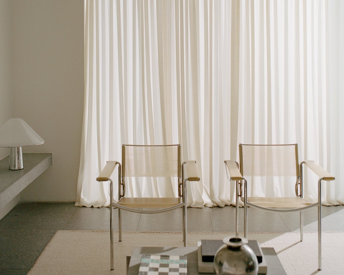 The Zero Basket rug in Sand Mix set in a bright living room, paired with a white set of curtains alongside two minimalistic armchairs and a coffee table made out of stone.