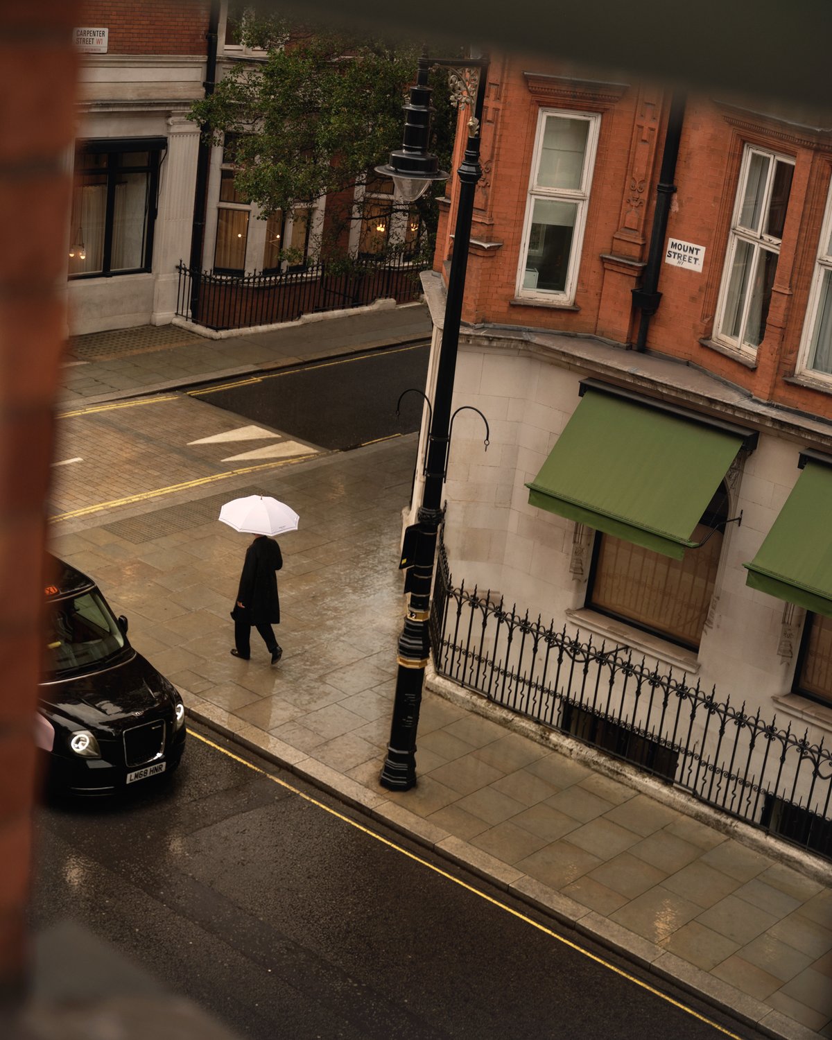 A view from the Nordic Knots store on Mount Street in London, showing a rainy street scene with a person walking under a white umbrella past red brick buildings and green awnings.