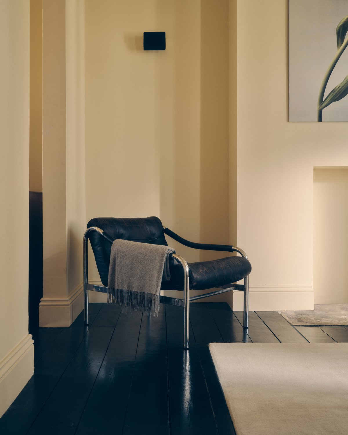 Tapis en laine moelleux Grand en Dusty White, photographié sur un sol en bois noir avec un fauteuil en cuir noir à côté.