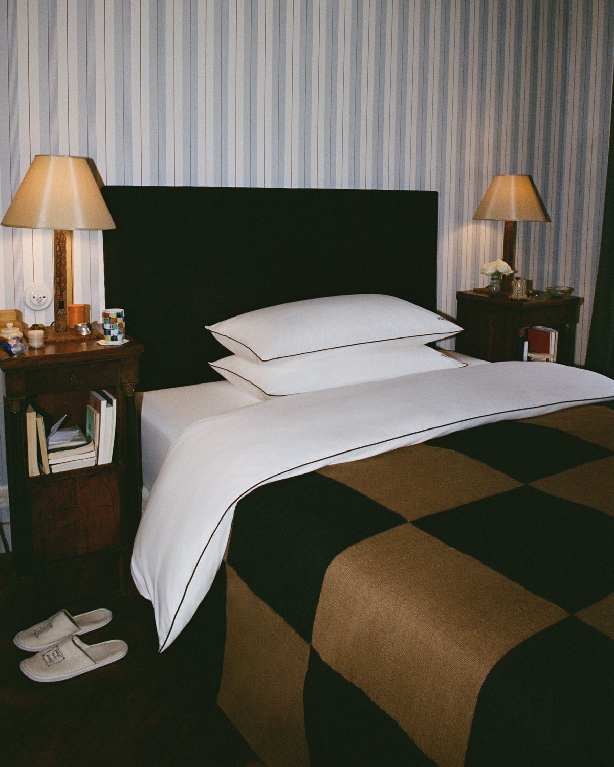 Der Square Kelim in Tobacco wird auf einem ordentlich gemachten Bett mit der Percale Duvet Cover in Crisp White/Black gezeigt, eingerahmt von zwei Holznachttischen mit passenden Lampen und einer gestreiften Tapete im Hintergrund.