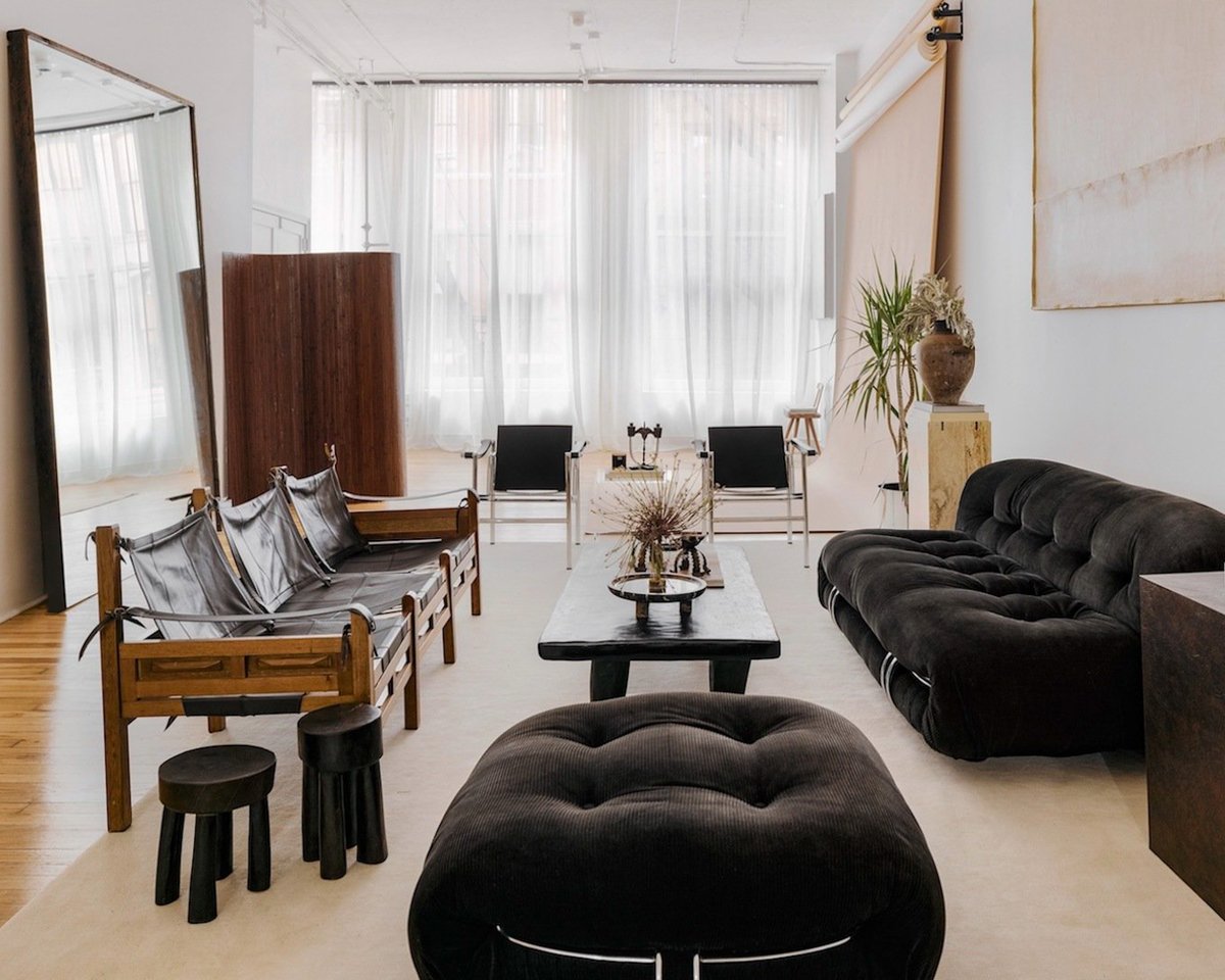 Flauschiger Wollteppich Grand in Dusty White, platziert in einem modernen Wohnzimmer mit künstlerischer Einrichtung, Holzsesseln und einer weichen schwarzen Sofagarnitur.