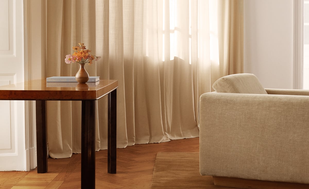 The Sheer curtains in Pale Sand are shown in a sunlit living room with a beige sofa, wooden table, and parquet floor, creating a soft and natural atmosphere.