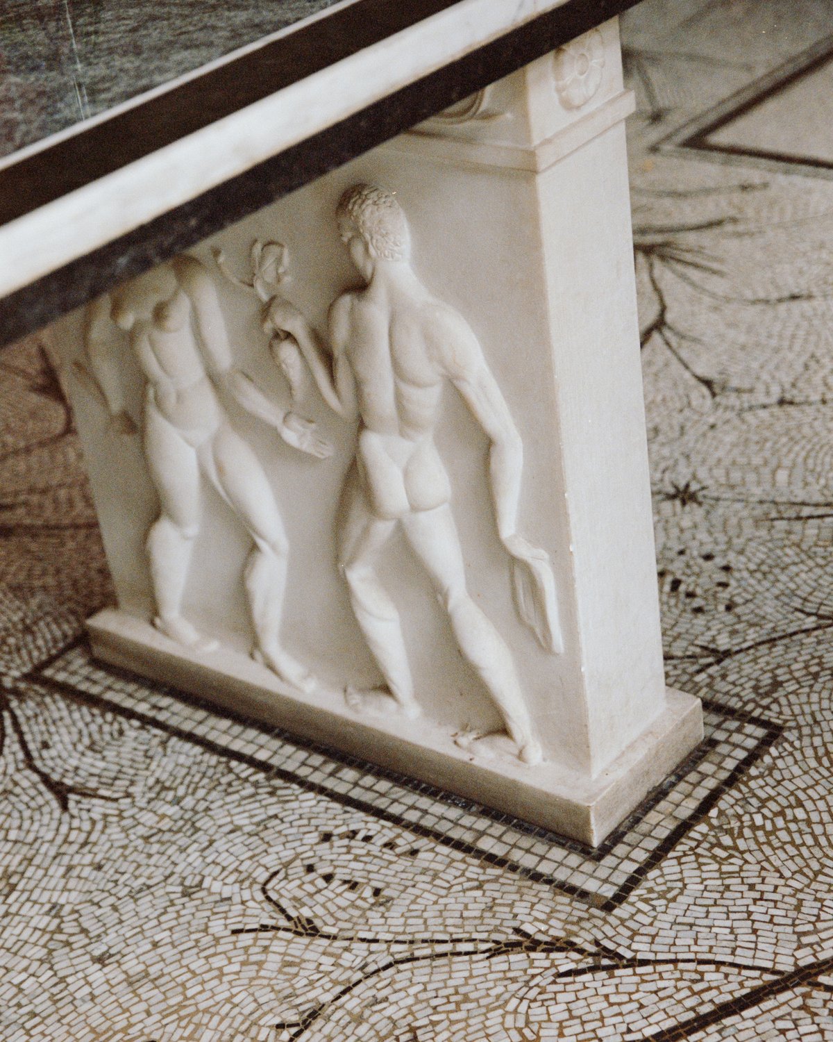 A marble table base featuring classical male and female figures in relief, set against a mosaic floor at Stockholm Concert Hall.
