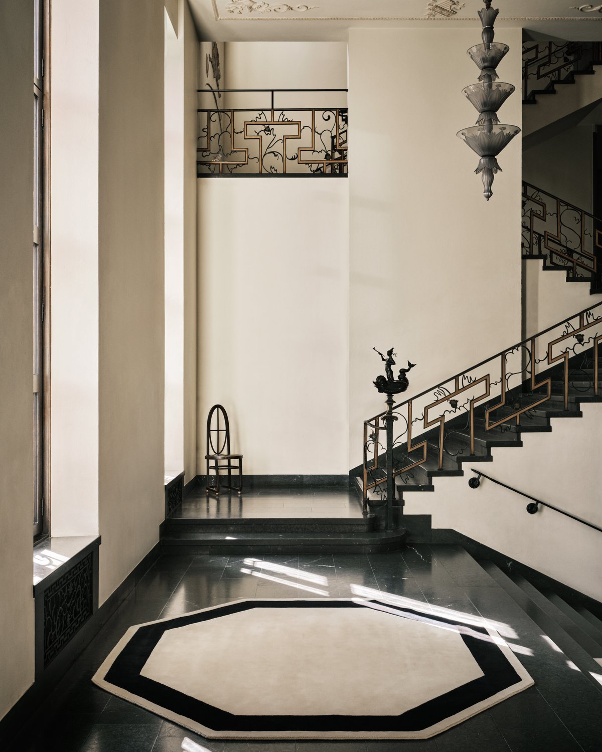 Grace 05 – Octagon — an octagonal rug in neutral beige framed by a bold black border, photographed inside Stockholm Concert Hall with sculptural stair railings and soft natural light.