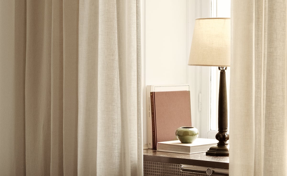 The Sheer Linen curtains in Almond are shown framing a softly lit window with a dark lamp, books, and a small ceramic bowl on the windowsill.