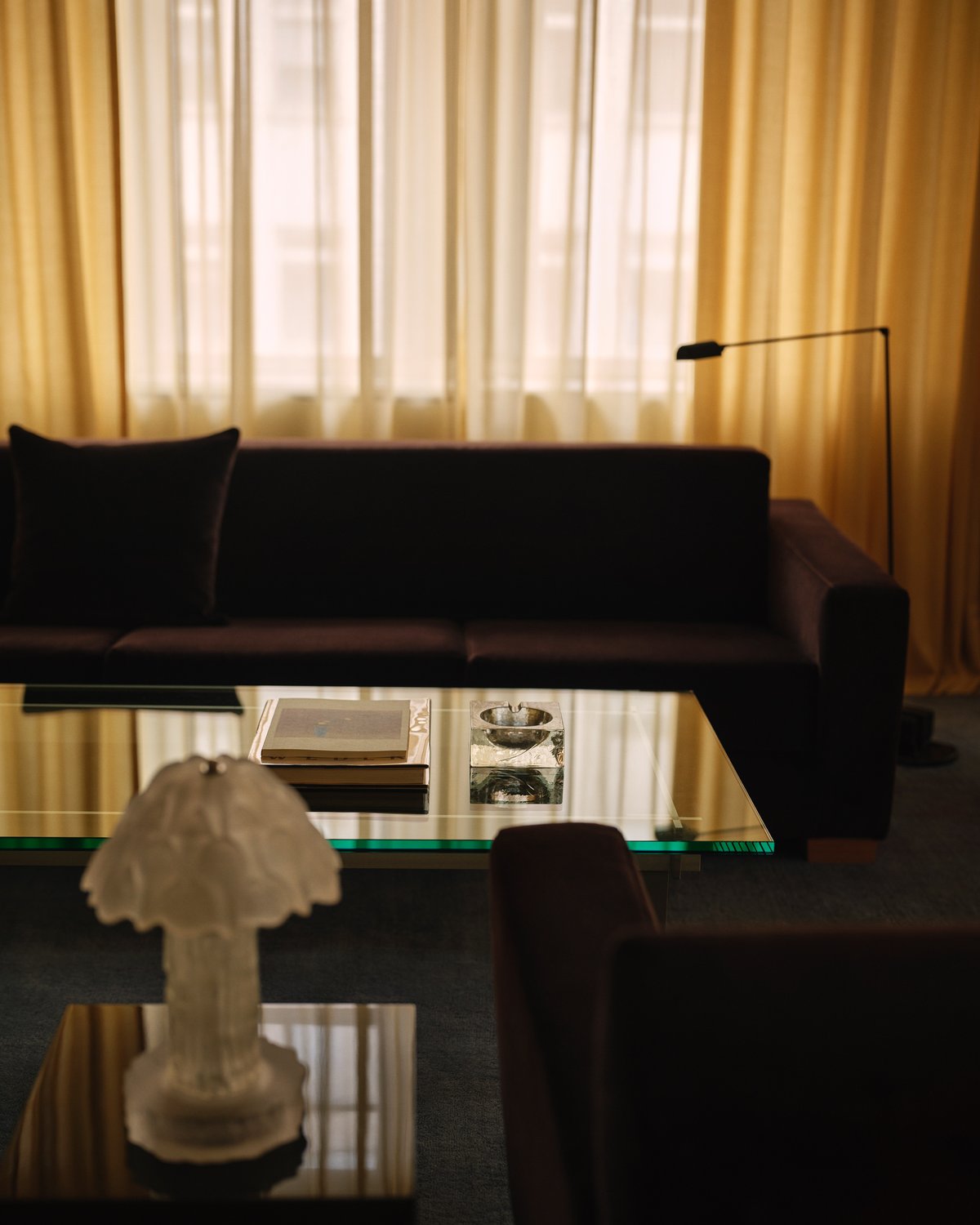 The Grand curtains in Leo and The Sheer curtains in Sunrise are shown in the Nordic Knots store on Mount Street in London, framing a softly lit lounge with dark velvet sofas and a glass coffee table, creating an elegant and cinematic atmosphere.