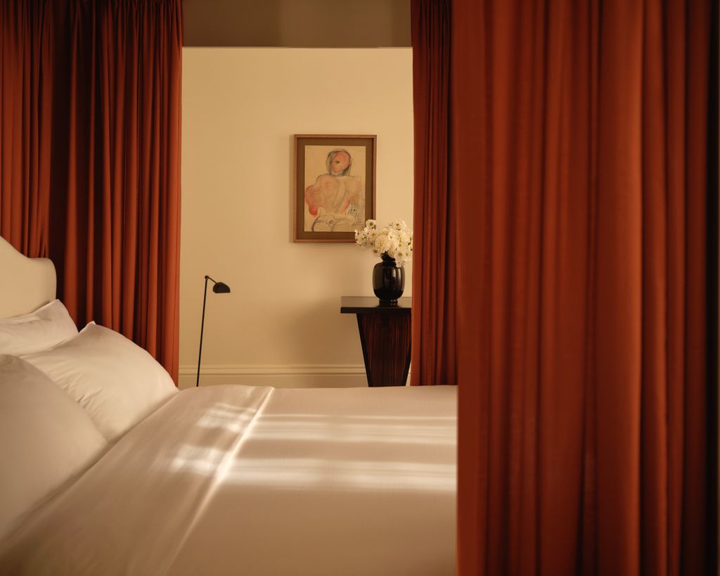 A calm bedroom setting in the Nordic Knots store on Mount Street, London. Crisp white bedding is framed by The Grand curtains in Burnt Red, with a dark wood side table, a floral arrangement, and an artwork adding warmth and balance to the scene.
