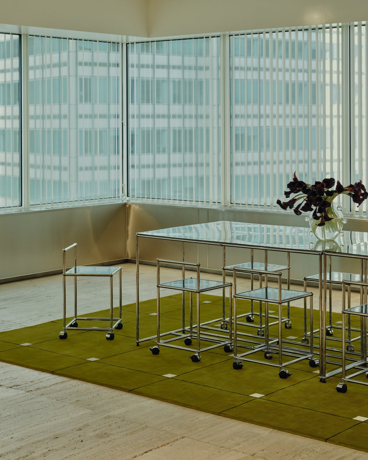 The plush wool rug Modern in Milano Green displayed in a bright dining room, paired with a glas dining table with metallic frame and matching chairs. On top of the table, there is a beautiful vase with burgundy flowers.