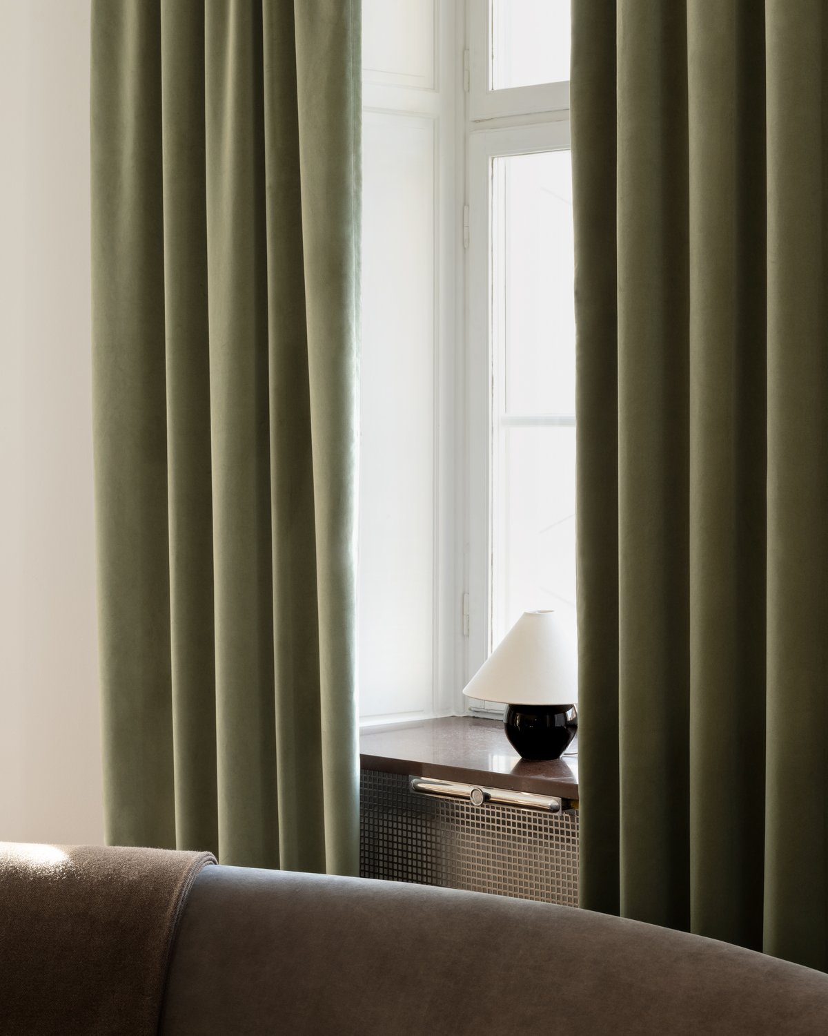 The Velvet Vorhänge in Olive Green, fotografiert im weichen Tageslicht, die ein Fenster mit einer kleinen Lampe einrahmen.