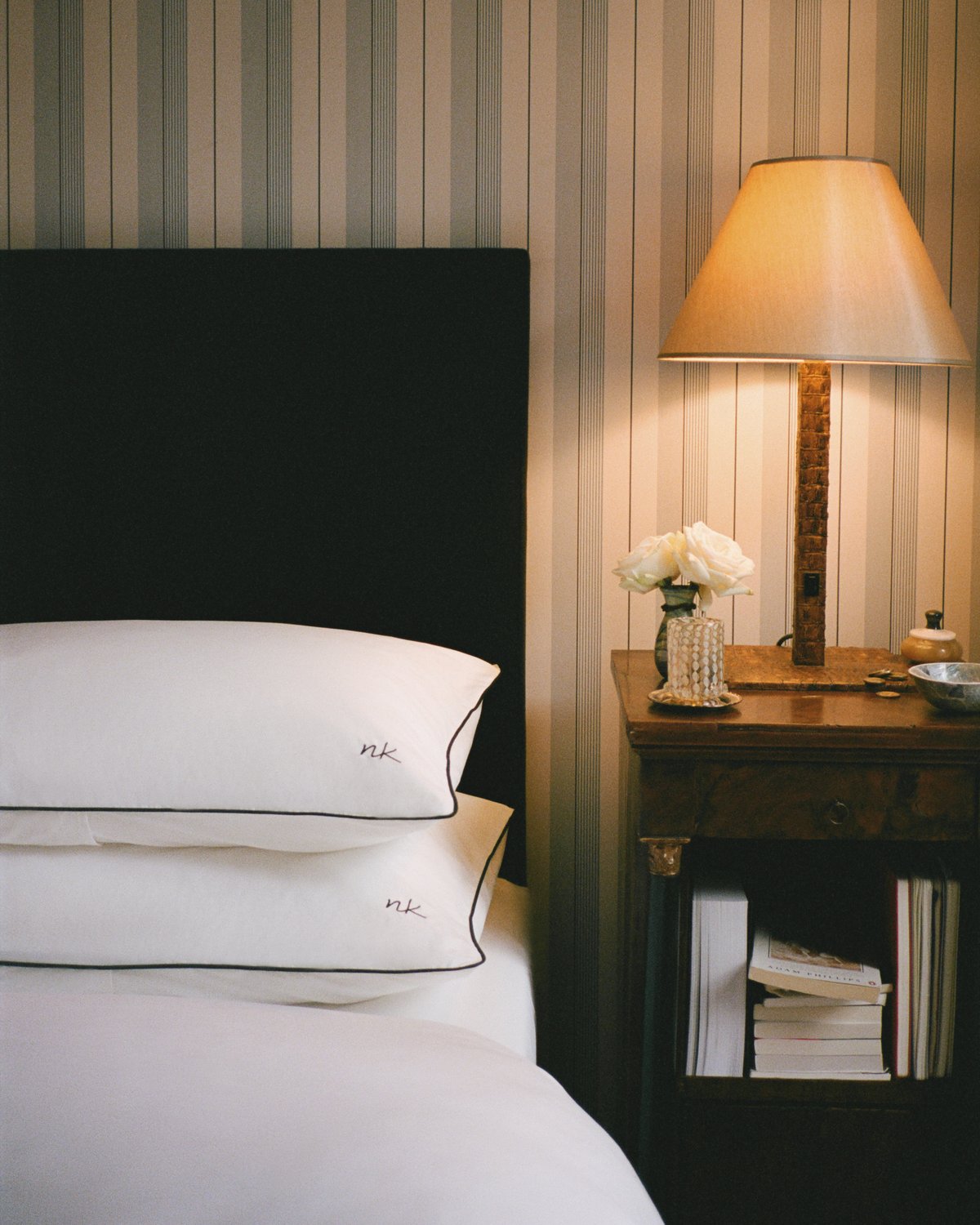 Die Percale Pillow Cases in Crisp White/Black sind ordentlich auf einem Bett neben einem Holznachttisch mit einer Lampe, Büchern und einer Vase mit weißen Blumen vor einer gestreiften Tapete gestapelt.