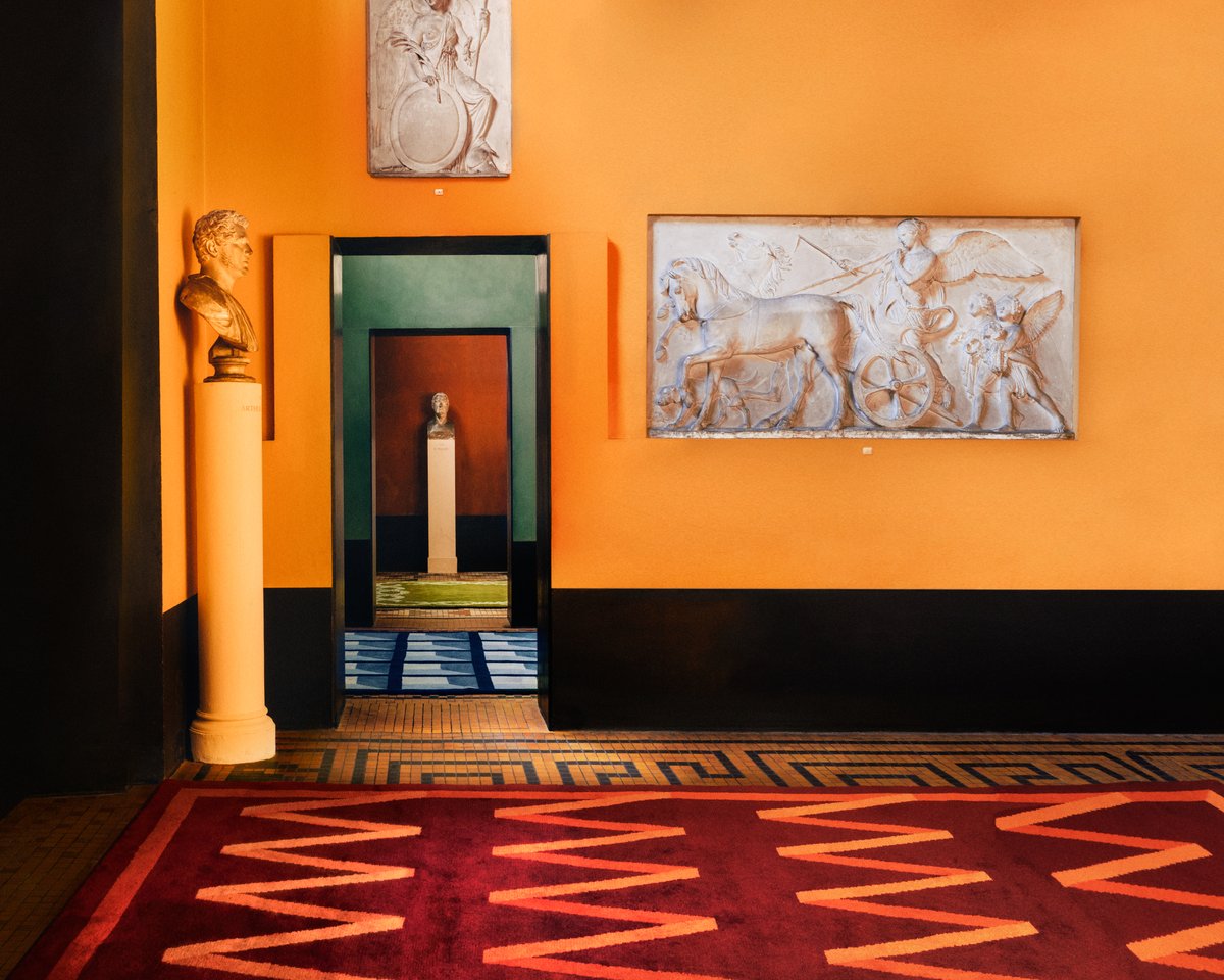 Den mjuka ullmattan Folding Ribbon - Burgundy från Campbell Reys kollektion 2023 fotograferad i ett färgstarkt rum med vacker konst på Thorvaldsens Museum i Danmark.