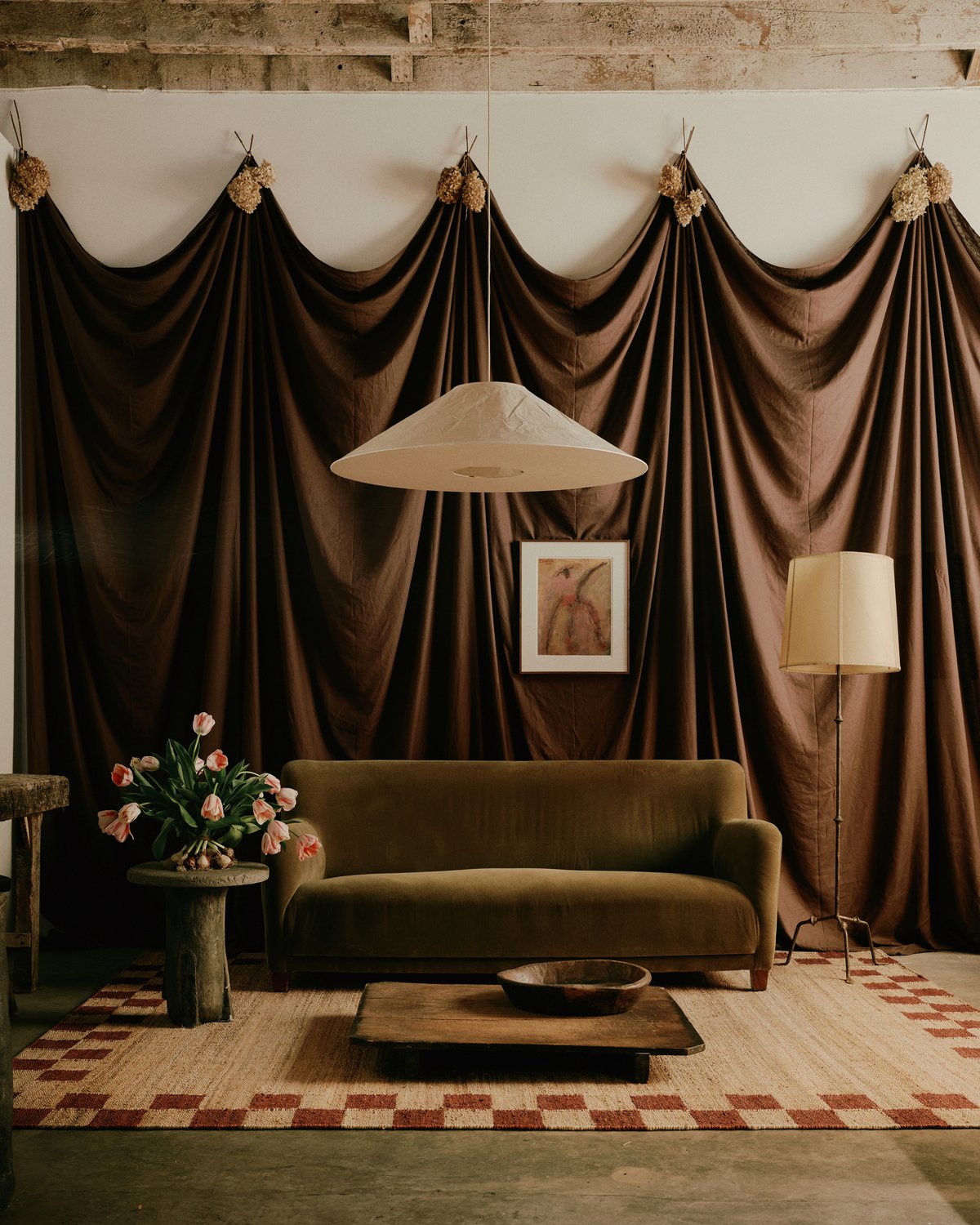 Warm-toned living room with dark draped curtains, an olive green velvet sofa, tulip flowers, and the Jute Border – Red rug with a checked border design.