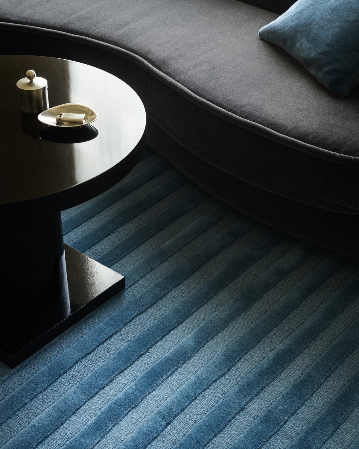 Park 03 rug in Steel Blue – a textured blue rug with tonal vertical stripes, anchoring a modern interior with a dark sculptural sofa and sleek black coffee table.