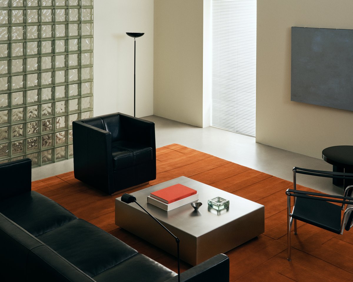 Plush wool rug Lux in Burnt Orange pictured in a brutalism style living room with black leather furniture and metallic coffee table.