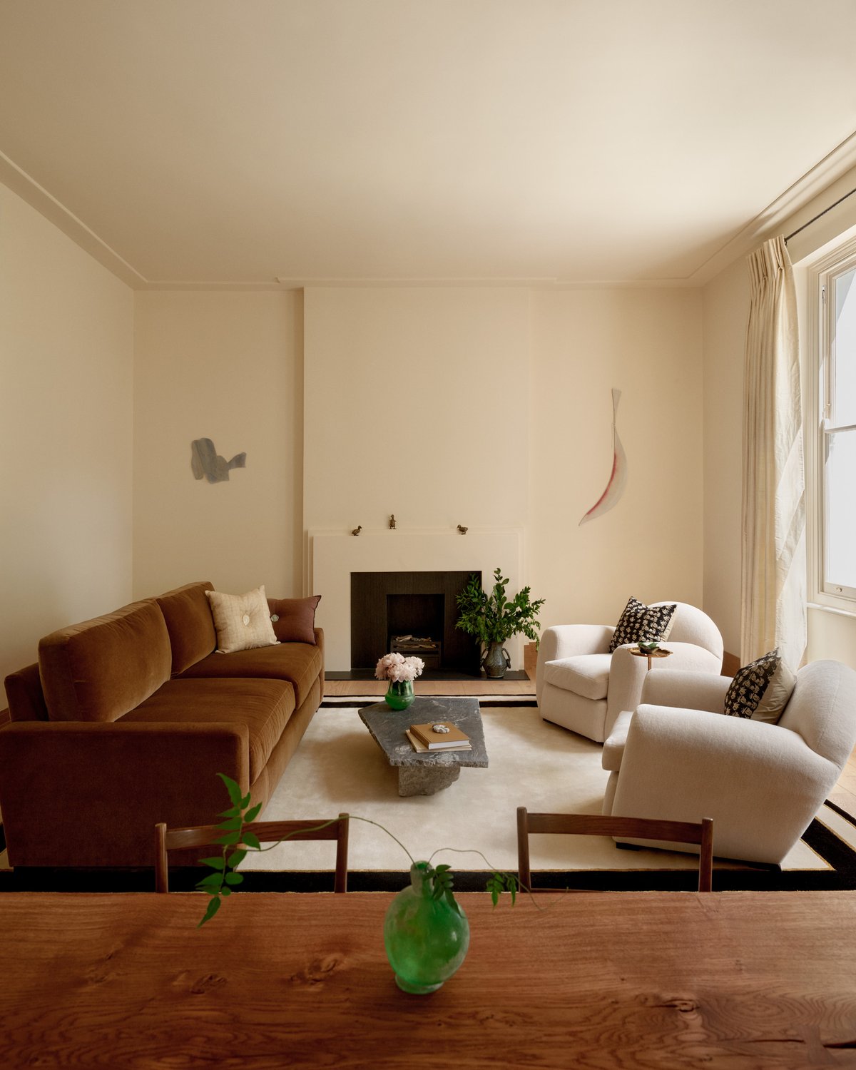 Gild 01 – Frame rug placed in a living room, featuring a soft beige surface with a subtle dark border, anchoring a seating area with a brown sofa, sculptural armchairs, and a low stone coffee table.