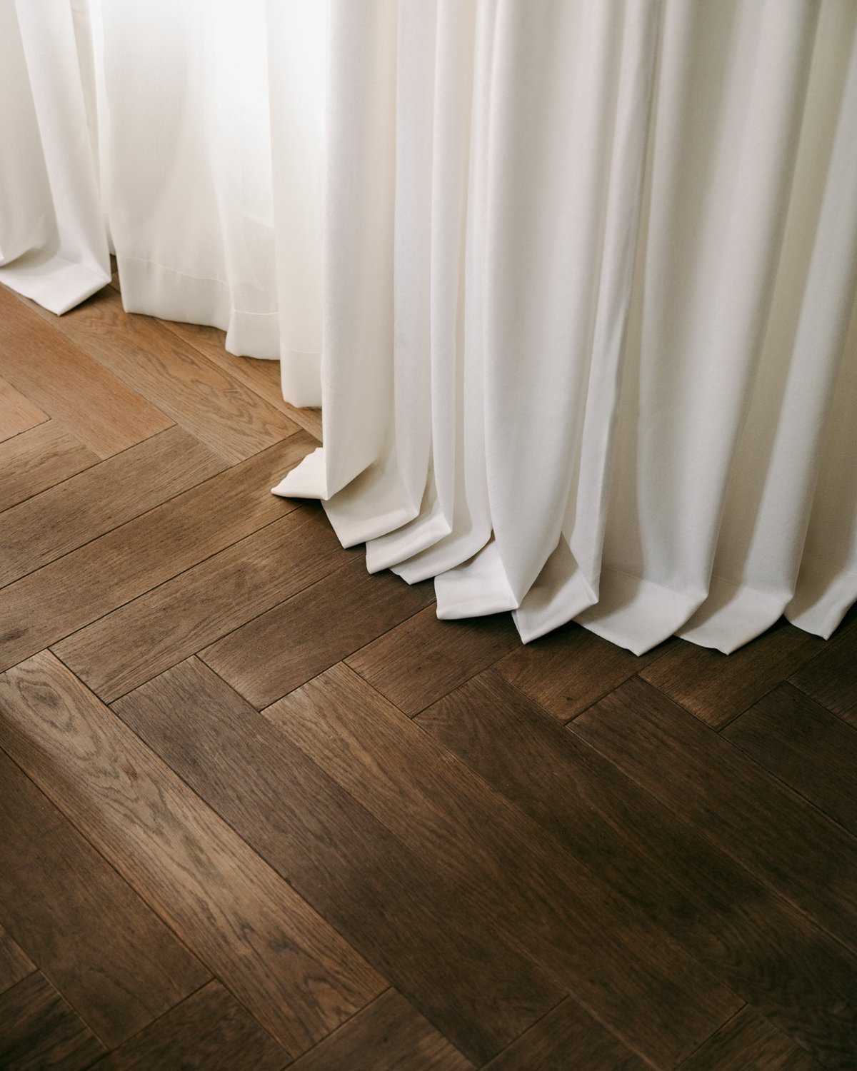 Close-up of The Grand curtain in Perfect White, showing a soft white fabric with a smooth woven texture and full-length folds resting on a wooden floor.