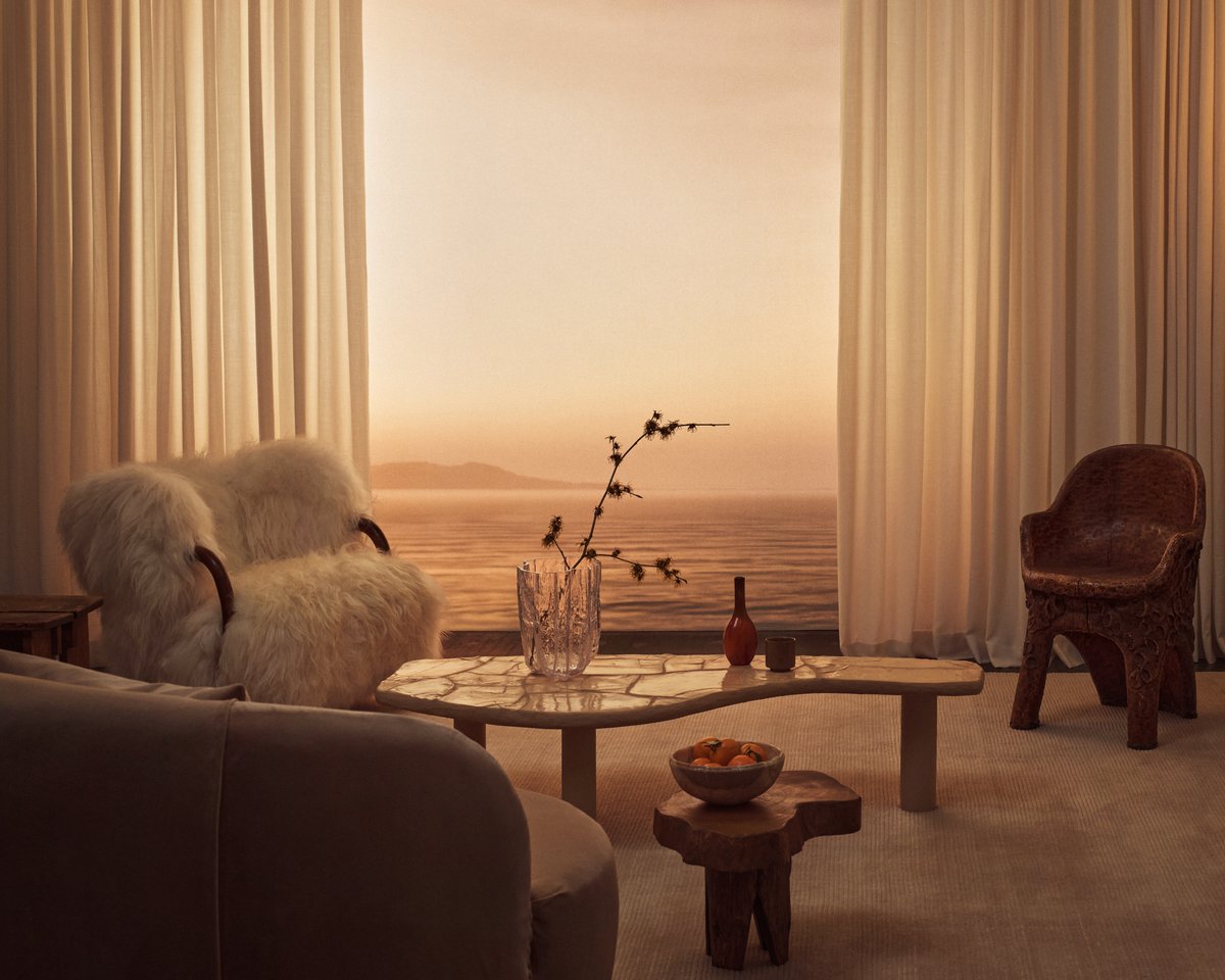 The Grand curtains in Perfect White pictured in a living room with neutral colored furniture and tall windows overlooking the sea.