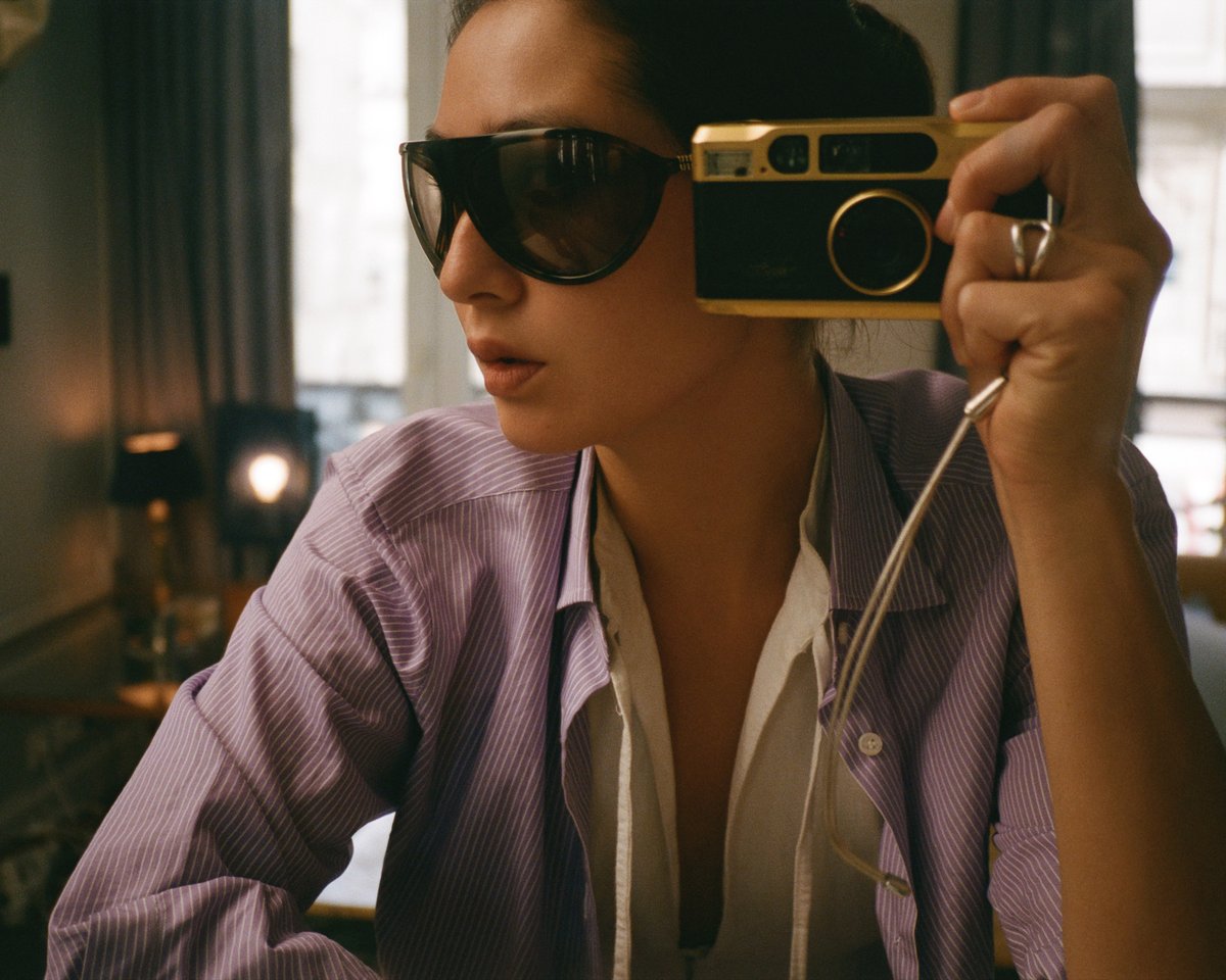 Diana Bartlett taking a self-portrait with a vintage-style camera, wearing dark sunglasses and a striped shirt in a softly lit interior with warm tones and elegant details.