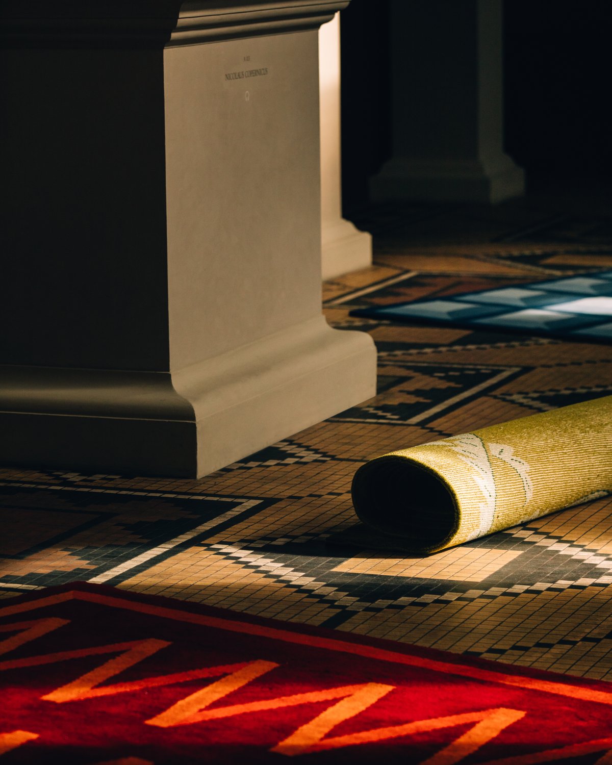 Närbild av den mjuka ullmattan Folding Ribbon - Burgundy från Campbell Reys kollektion 2023, fotograferad på ett färgstarkt mönstrat golv på Thorvaldsens Museum i Danmark.