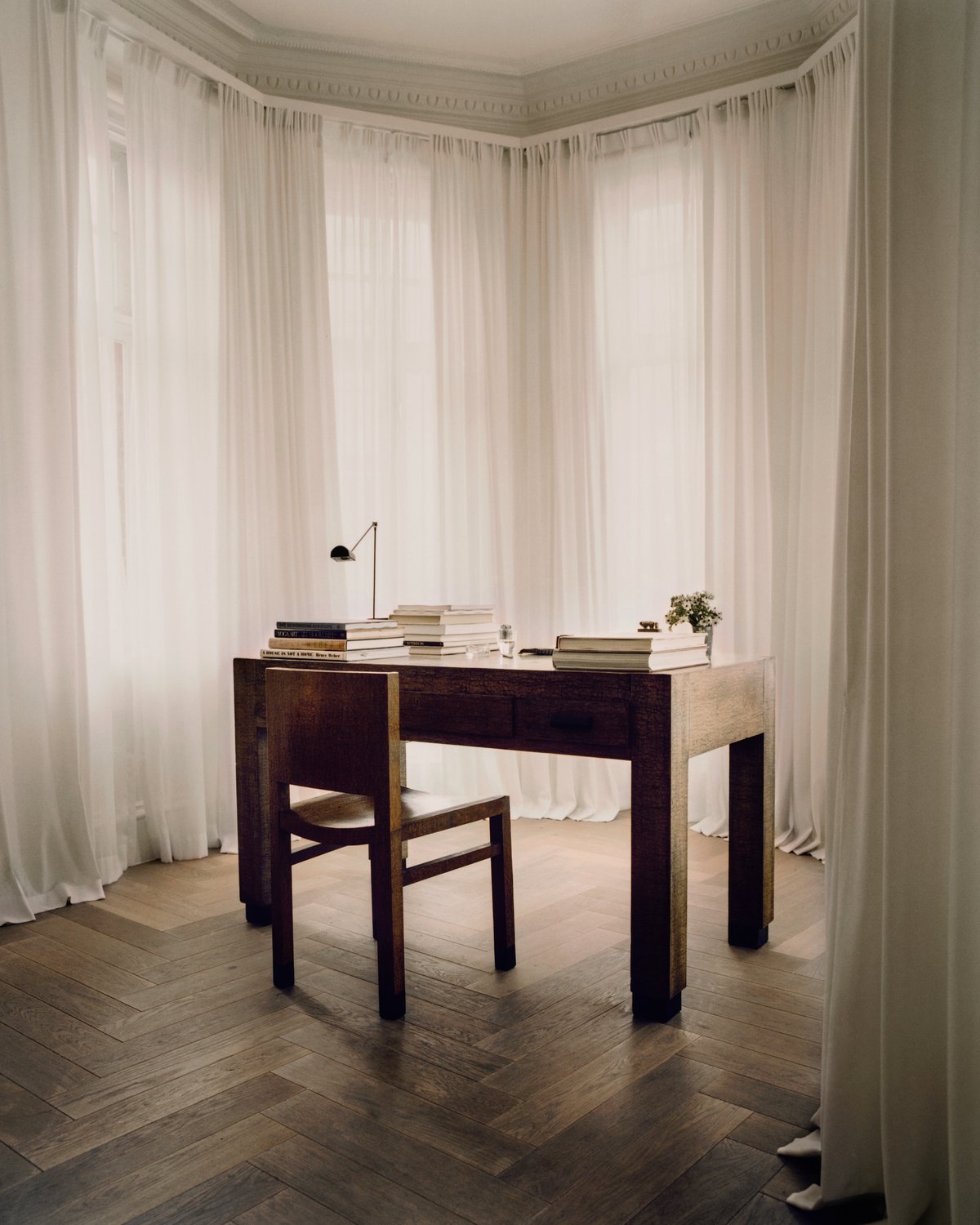 The Grand curtain in Perfect White styled in a room, showing a soft white fabric with a smooth woven texture and full-length vertical folds filtering light around a wooden desk and chair.