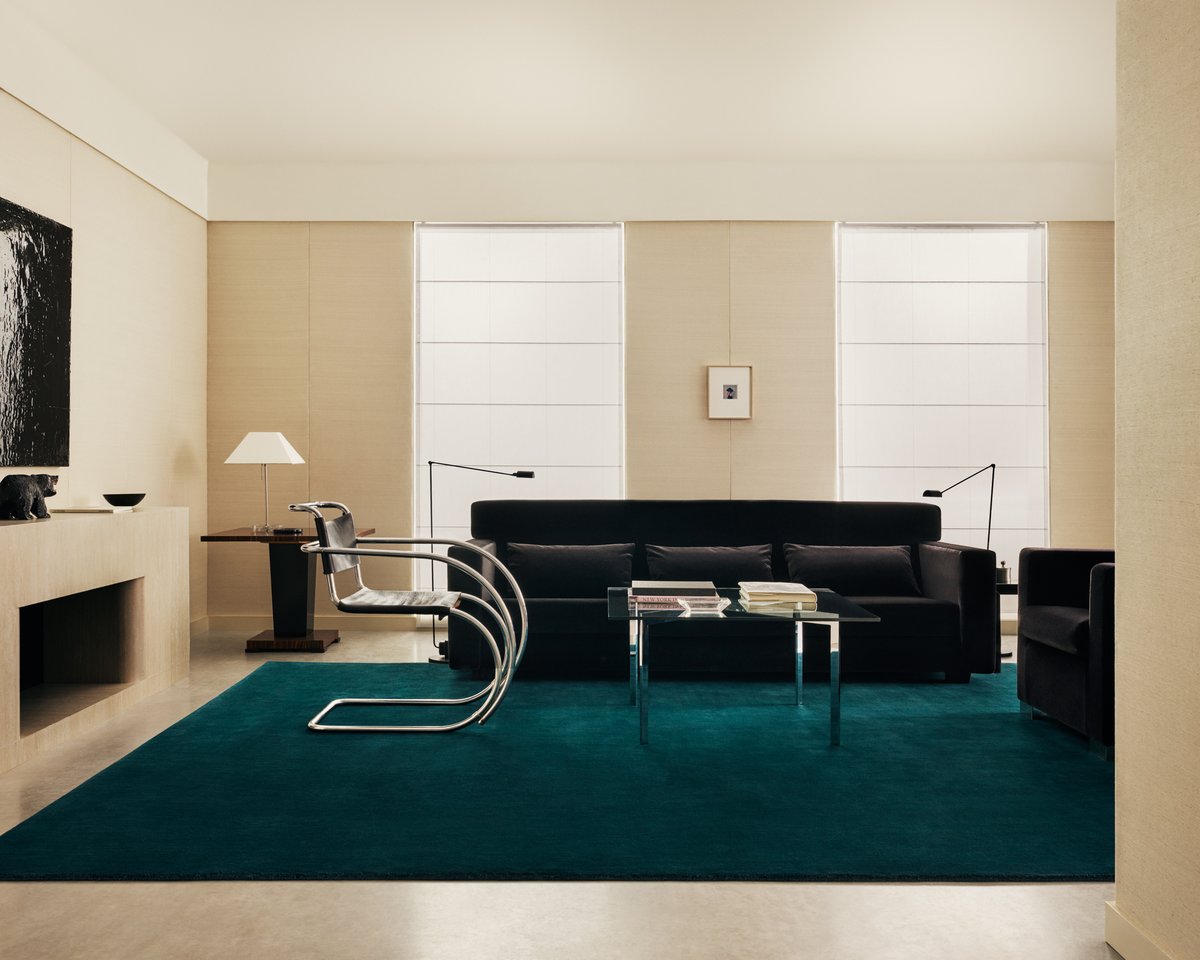 Wide in-room shot of the Grand rug in Emerald Green, grounding a black sofa and glass coffee table in a calm, minimal living space.