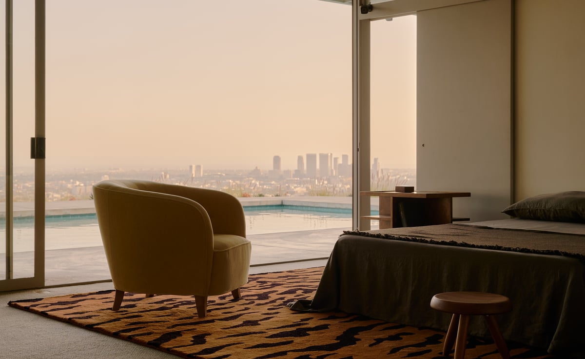 Tigris – Rust/Black brings bold contrast and warmth to this serene, modern bedroom. Its striking tiger-stripe pattern anchors the space beneath a mustard-toned armchair and charcoal-toned bed. Floor-to-ceiling windows open to a panoramic city view and pool, while the soft light and natural textures create a calm, curated atmosphere.