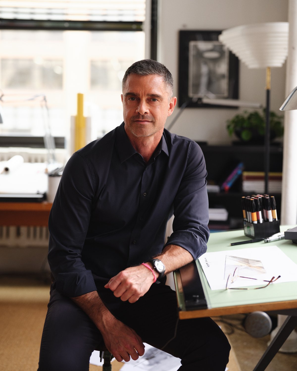 A designer seated in a studio workspace in Soho, New York, surrounded by sketches, tools, and material samples.