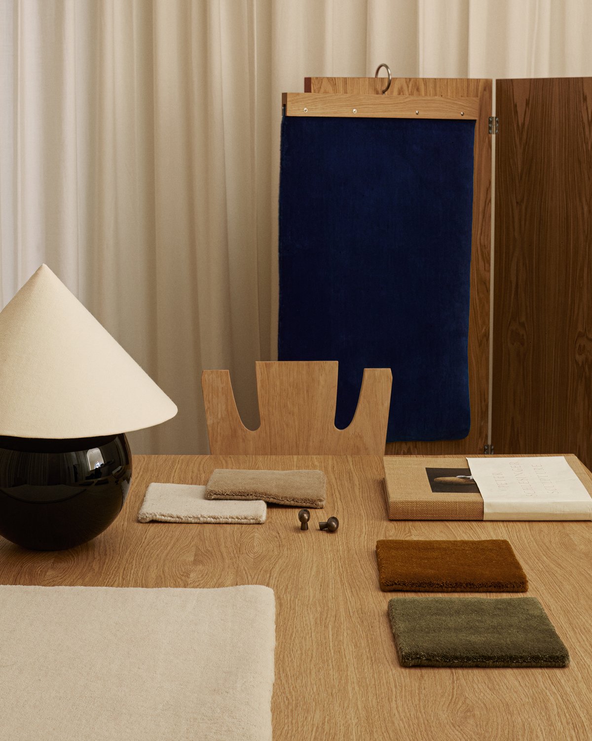 Muster des Plüschwollteppichs Grand in verschiedenen Farben, abgebildet auf einem Holztisch mit moderner Lampe und Coffeetable-Buch.