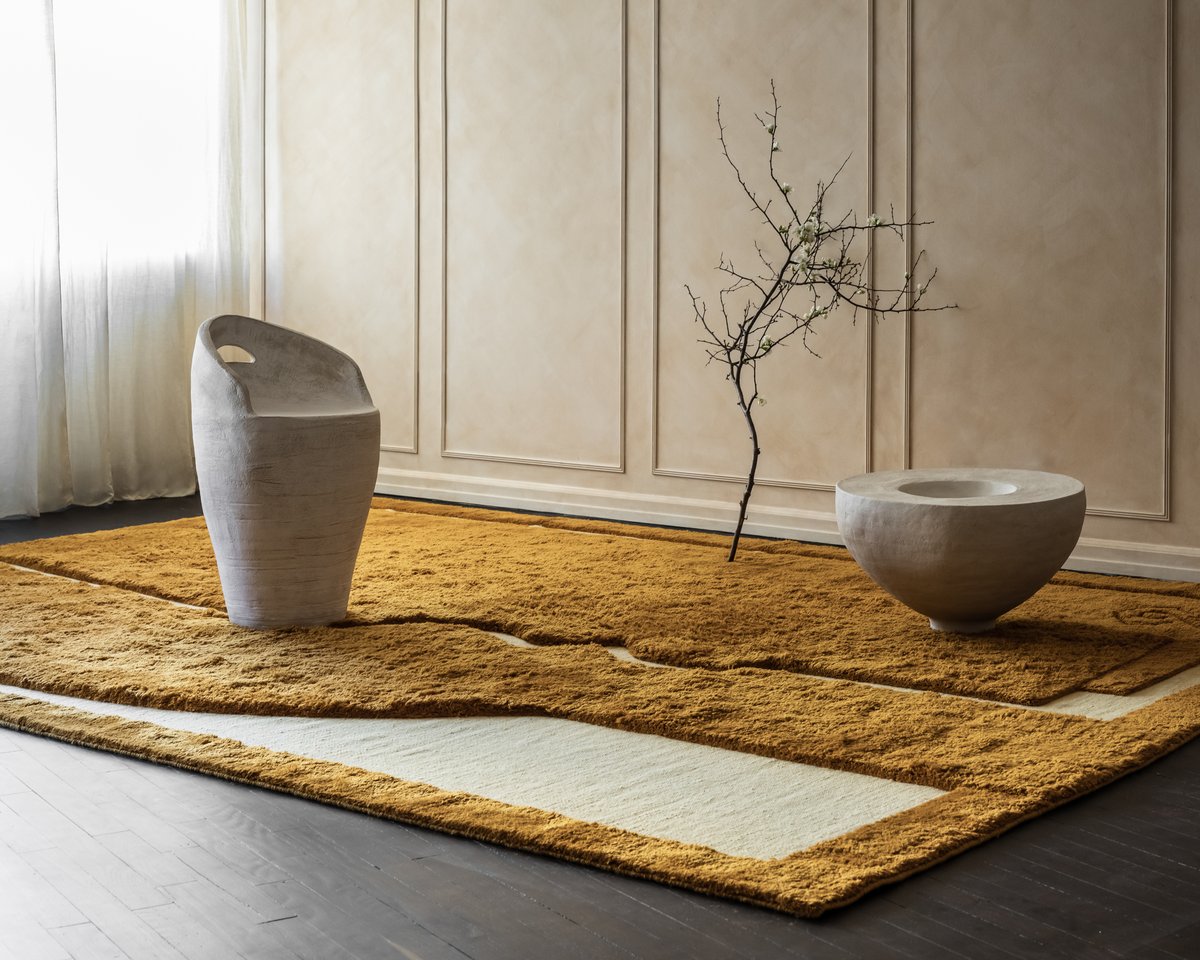 Face rug in Warm Ochre pictured with sculptural stone elements, a tree branch, and white curtains in the background.