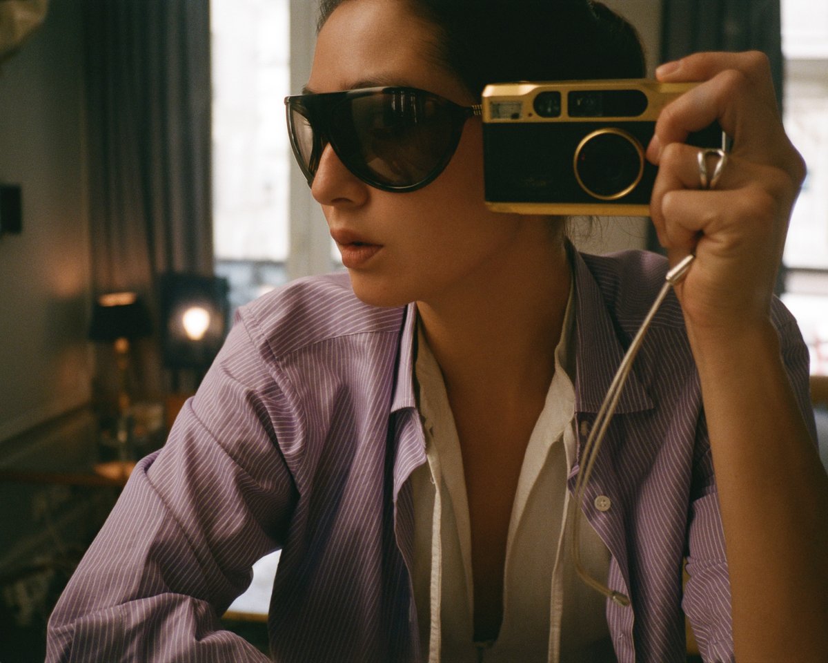 Diana Bartlett macht ein Selbstporträt mit einer Vintage-Kamera, trägt dunkle Sonnenbrillen und ein gestreiftes Hemd, in einem sanft beleuchteten Raum mit warmen Tönen und eleganten Details.