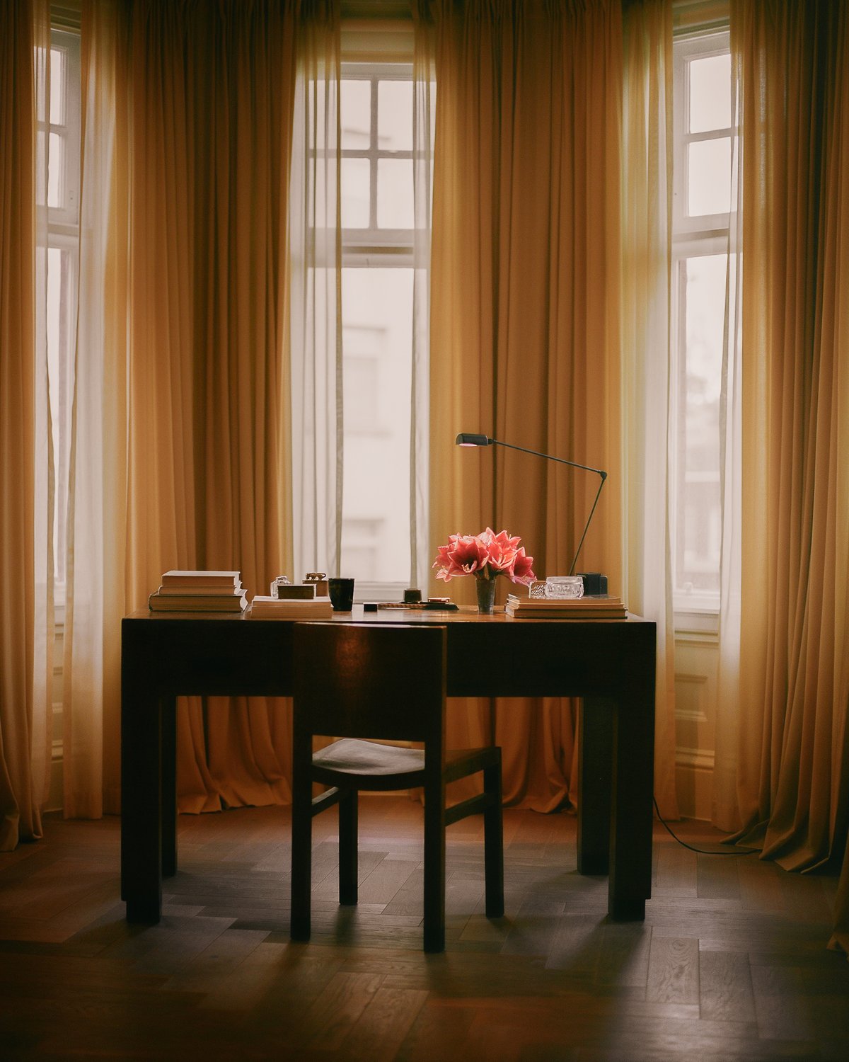 The Grand curtain in Leo styled around a window behind a writing desk, showing a warm golden ochre fabric with a smooth woven texture and full-length vertical folds filtering light.