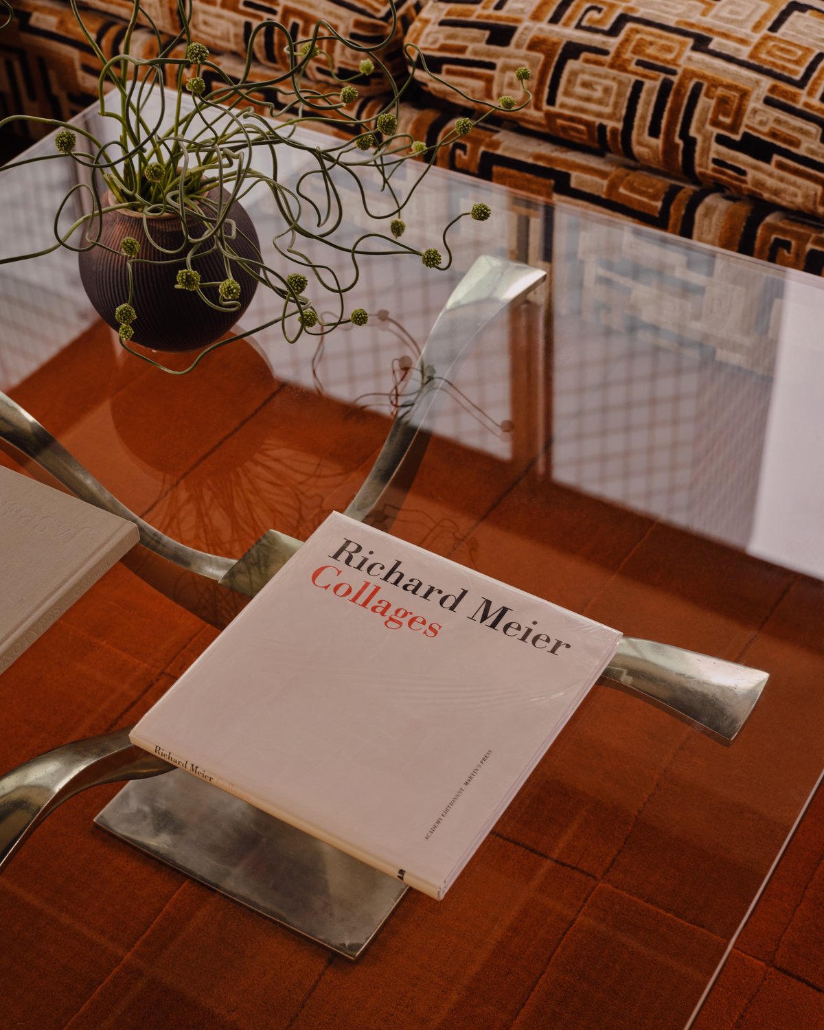 Le tapis Lux 2 en Burnt Orange placé sous une table en verre sculpturale avec un cadre métallique, à côté d’un canapé à motifs bruns.