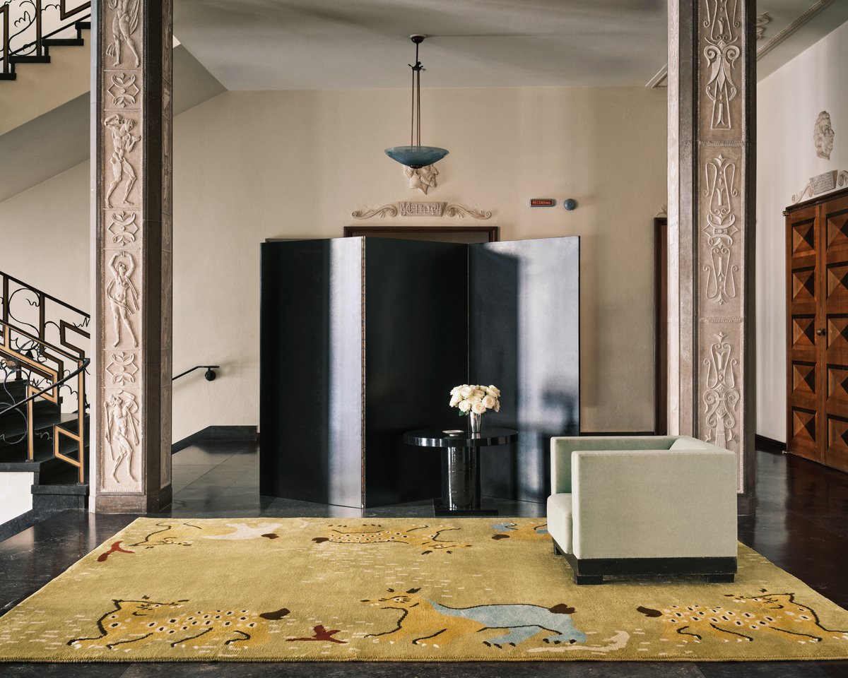 Grace 03 – Fauna — styled inside Stockholm Concert Hall. A golden beige rug with playful animal forms and organic patterns, paired with sculptural furniture and neoclassical details.