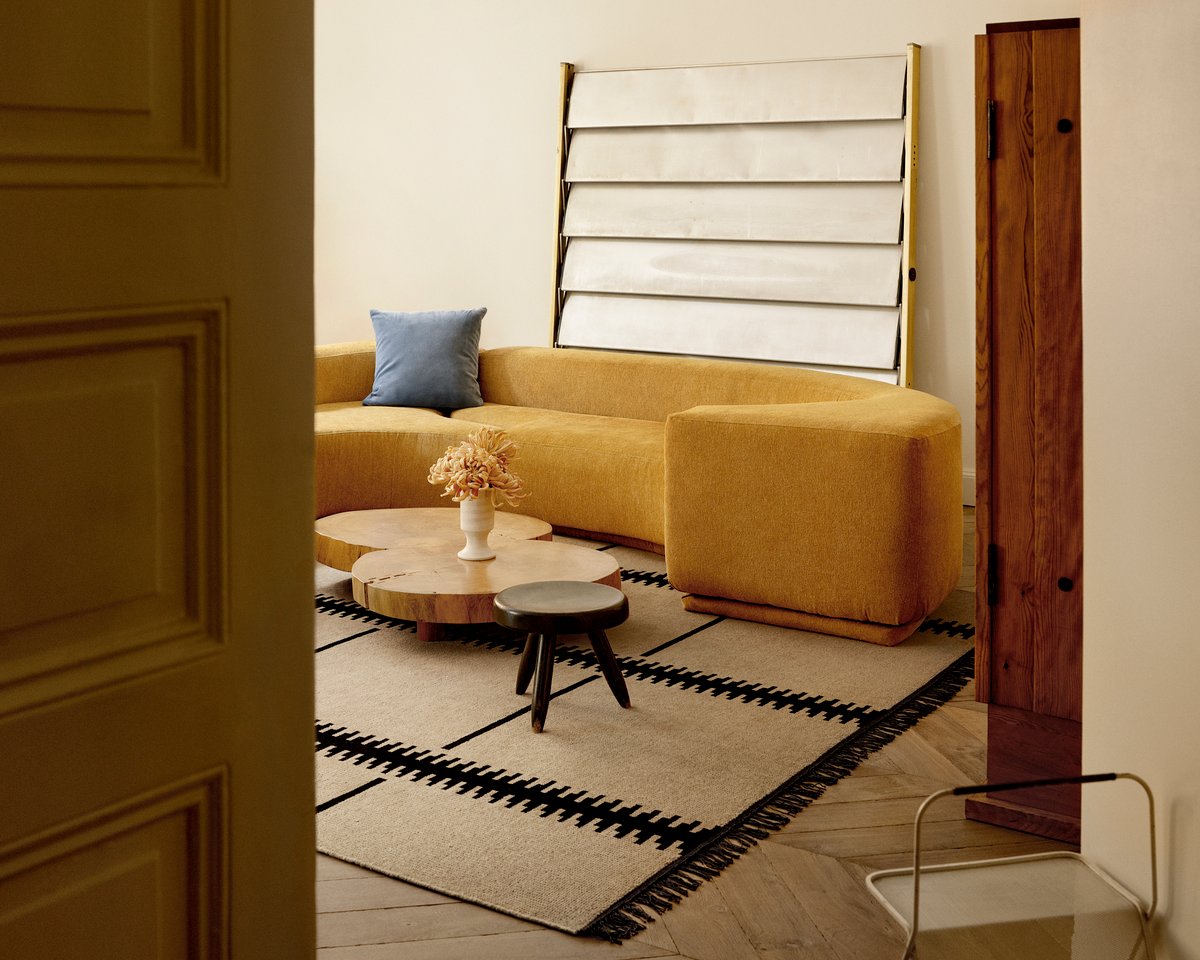 Warm living room with a curved mustard sofa, round wooden coffee tables, and the Elder 06 – Black rug featuring a bold linear border pattern for a modern graphic touch.