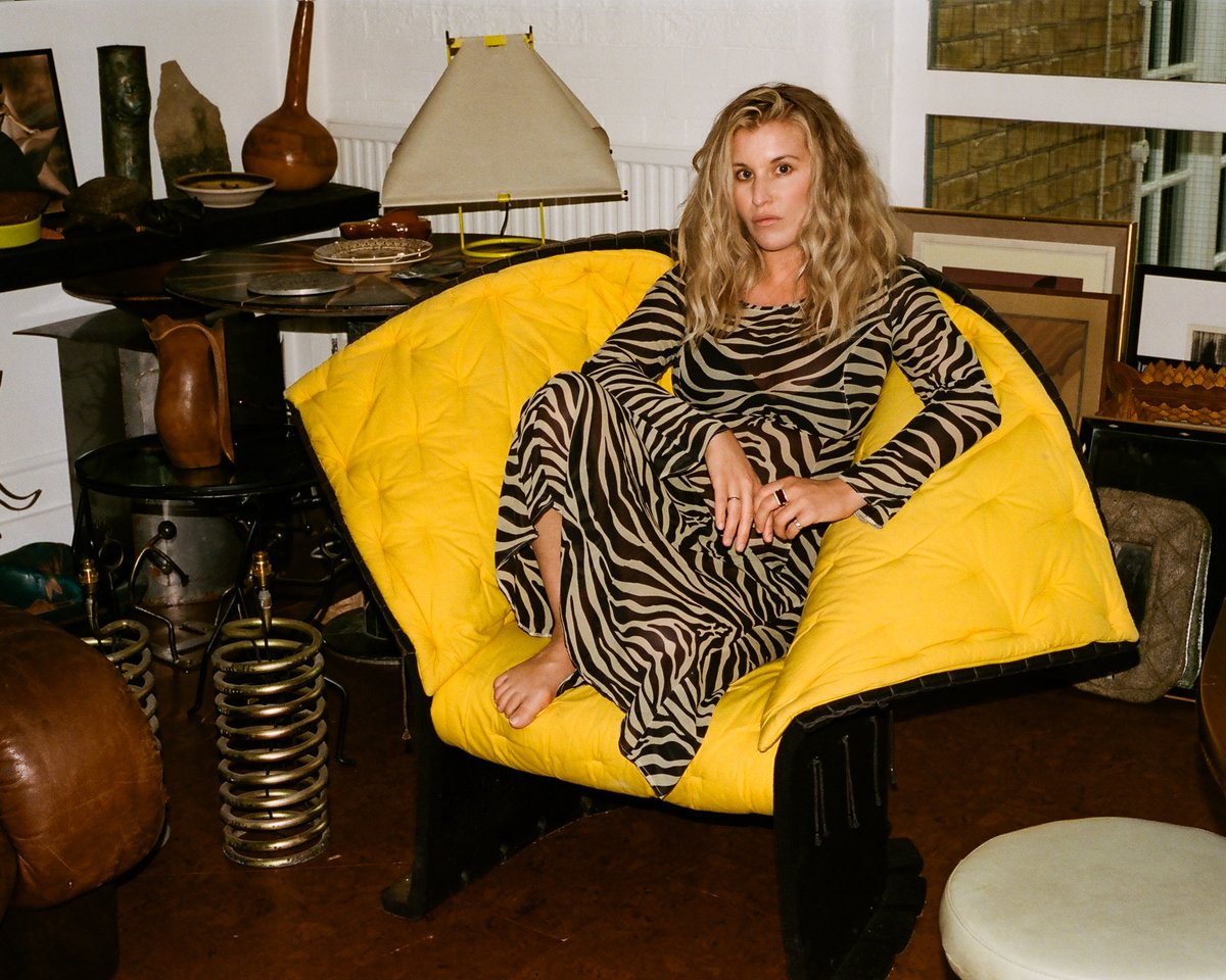 Hollie Bowden fotografiert in einem leuchtend gelben Sessel sitzend vor einem Tisch voller Einrichtungsgegenstände und Kunstwerke.