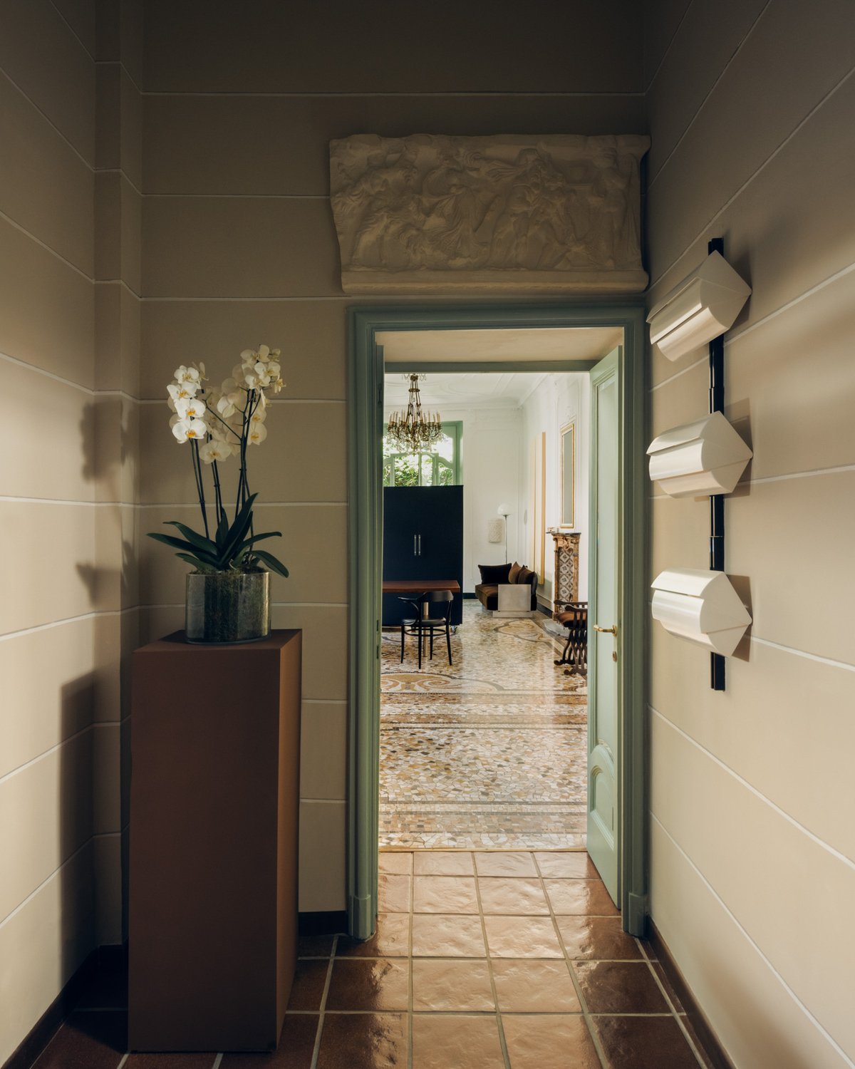 Ein schmaler Flur mit Fliesenboden und einem salbeigrünen Türrahmen, der in einen hellen Raum mit Terrazzoboden, Kronleuchter und skulpturalen Möbeln führt.