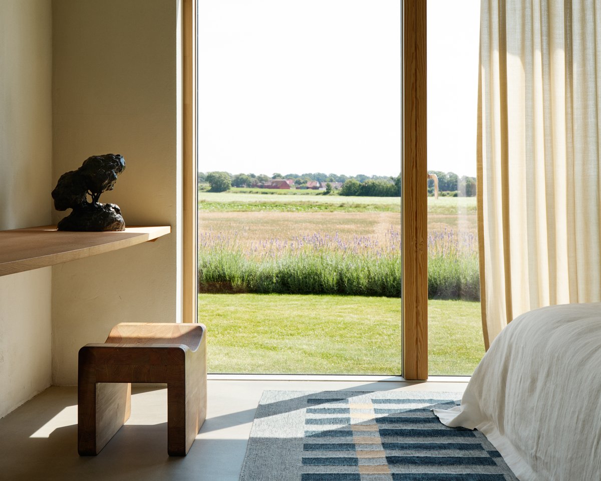 A bright bedroom with Tradition 01 in Blue, under a bed with white linens, next to a desk with a wooden stool, framed by natural curtains and meadow views.