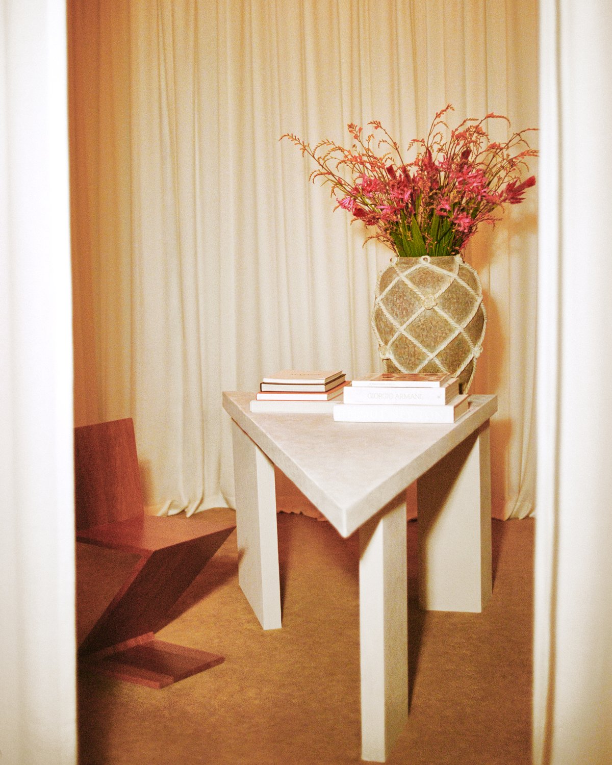 The Grand curtains in Perfect White depicted behind a large ceramic flower vase on top of a white, triangular office table next to a modern wooden chair resting on plush wool rug Grand in Leo in the Stockholm Flagship store upstairs curtain room.